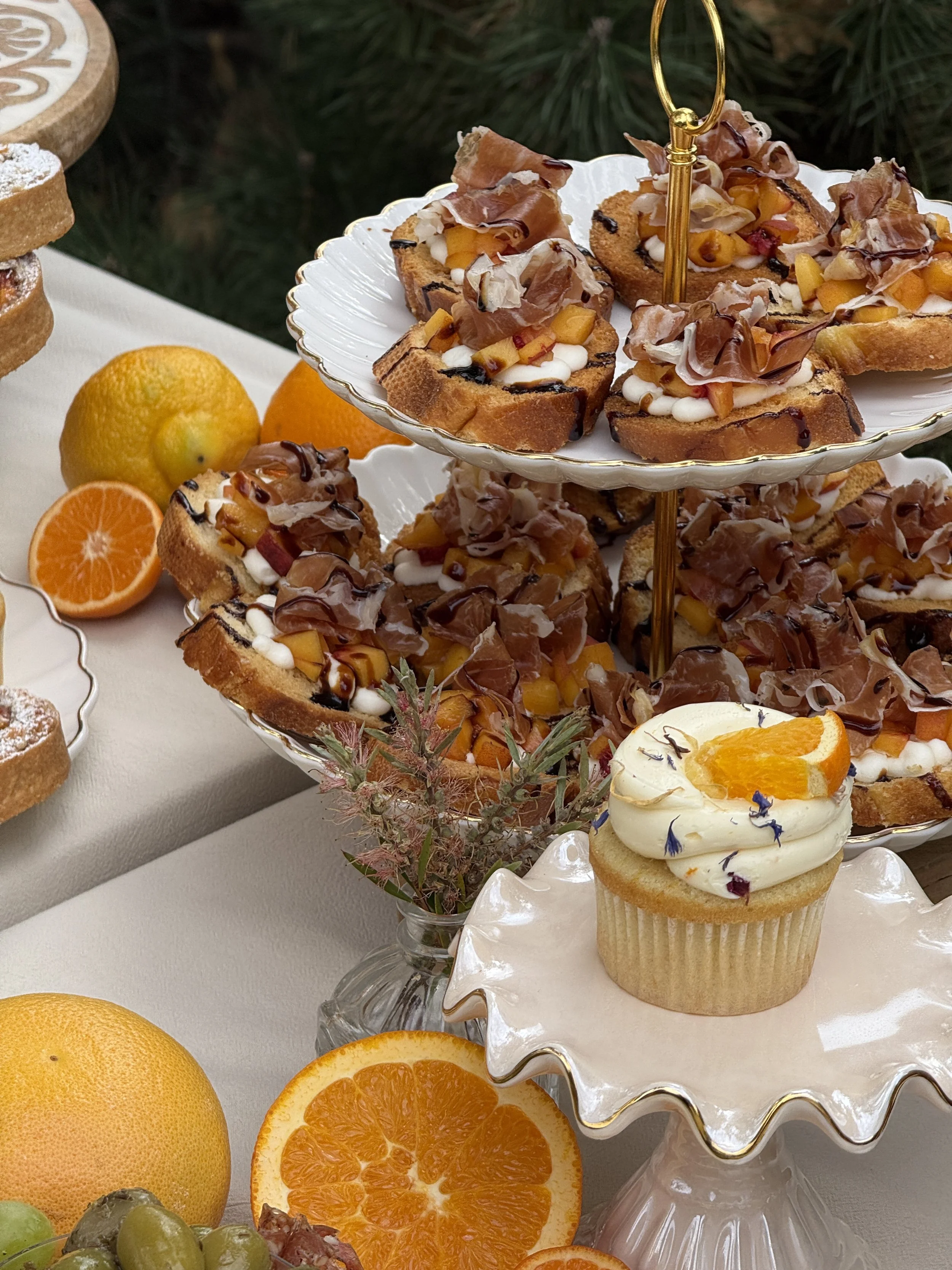 Assorted fruit-topped desserts including a cupcake with orange slices, on a decorated cake stand with oranges, grapes, and other fruit around, against a backdrop of greenery.