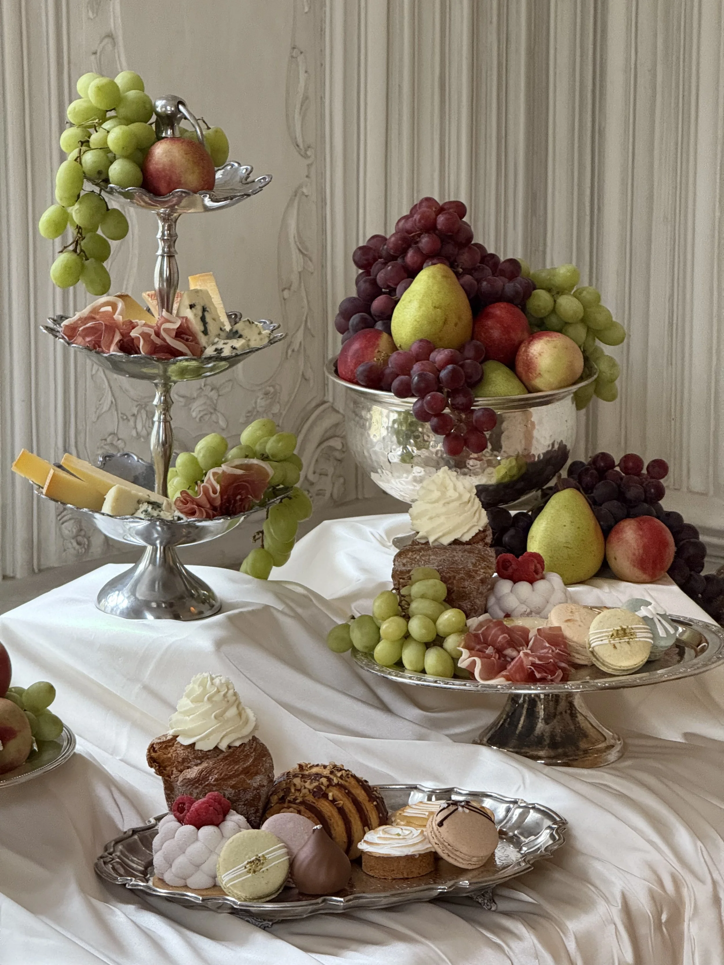 A tiered tray with grapes, cheese, and prosciutto, and a large glass bowl of assorted grapes and pears surrounded by desserts like macarons, cake, and cookies on a white tablecloth.