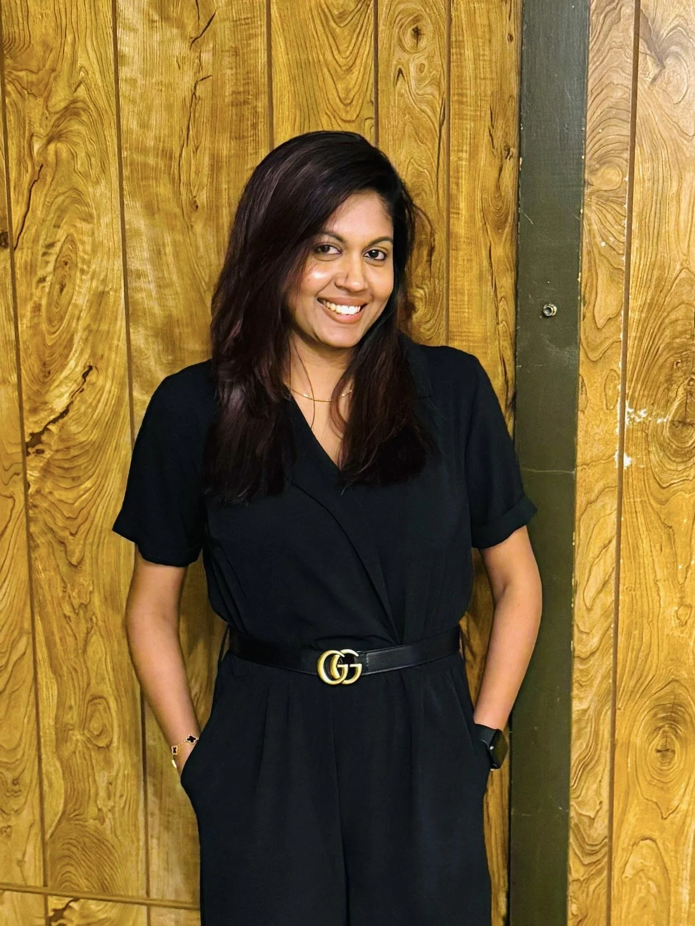 A woman with dark hair smiling, wearing a black outfit with a Gucci belt, standing in front of wooden panels.