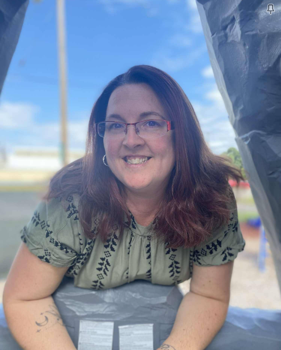 Woman with glasses and glasses with reddish-brown hair smiling outdoors in front of a blue sky with some clouds, wearing a patterned blouse.