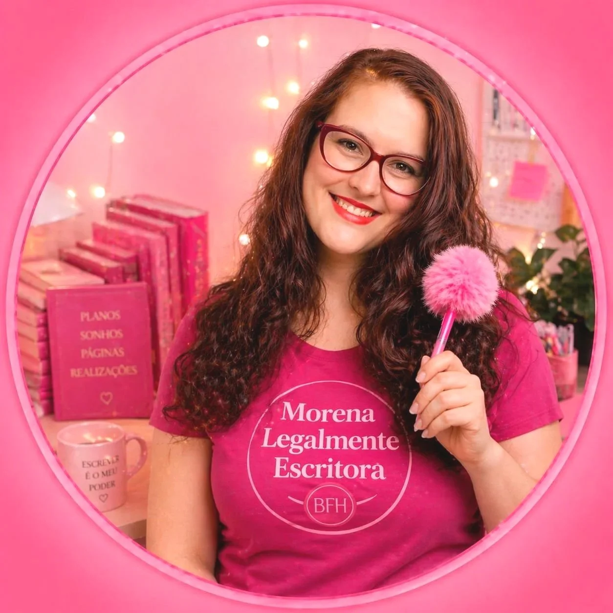 Mulher com cabelo cacheado e óculos de armação vermelha, sorrindo, segurando um funil de pelúcia rosa, usando uma camiseta rosa com frase 'Morena Legalmente Escrita'. Ao fundo, livros cor de rosa e uma caneca com mensagens e uma vela acesa, em um ambiente decorado com luzes brancas e plantas, tudo com tema rosa.
