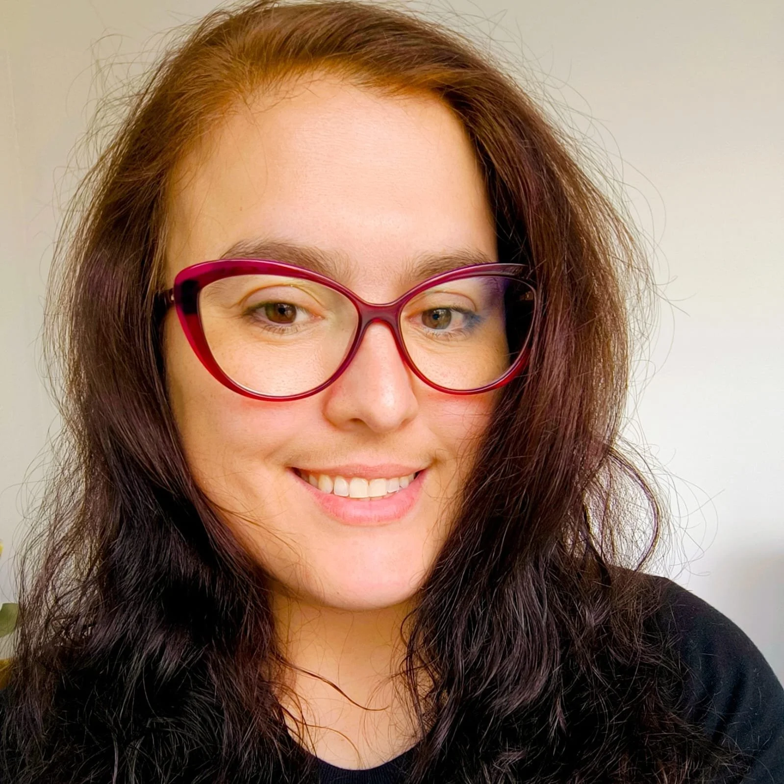 Retrato de uma mulher com óculos vermelhos, cabelos castanhos e sorriso, em um fundo branco.