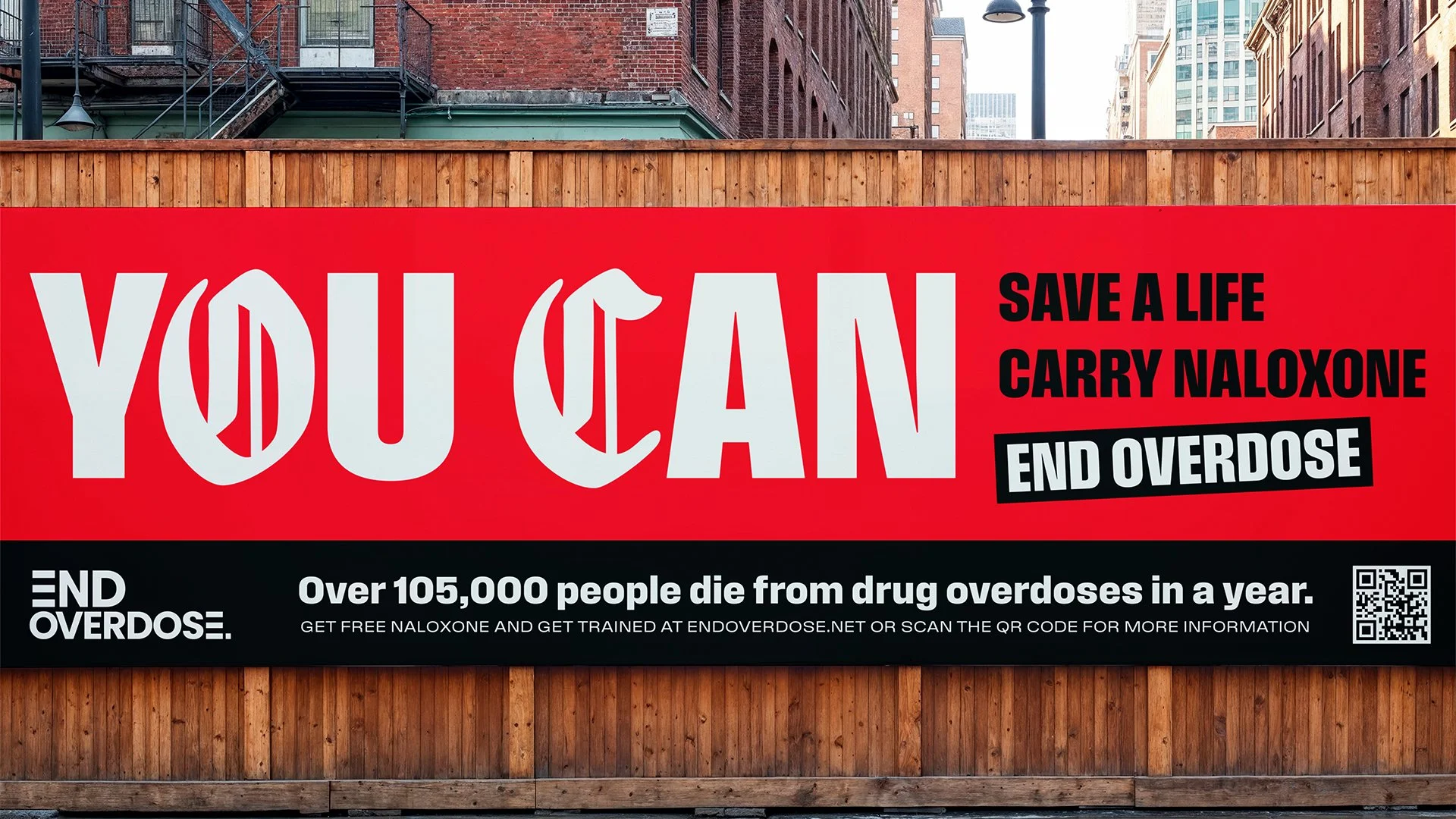 A red billboard with bold white text that reads "YOU CAN" and smaller black text that promotes saving lives by carrying Naloxone to prevent drug overdoses. The billboard includes a QR code and a message about overdose statistics, set against an urban city background with brick buildings and wooden fencing.