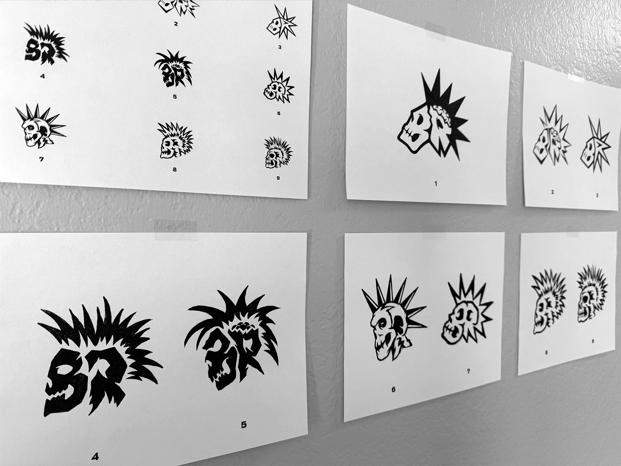 Black and white sheets of art illustrations featuring skulls, faces, and spiked designs, arranged on a wall.