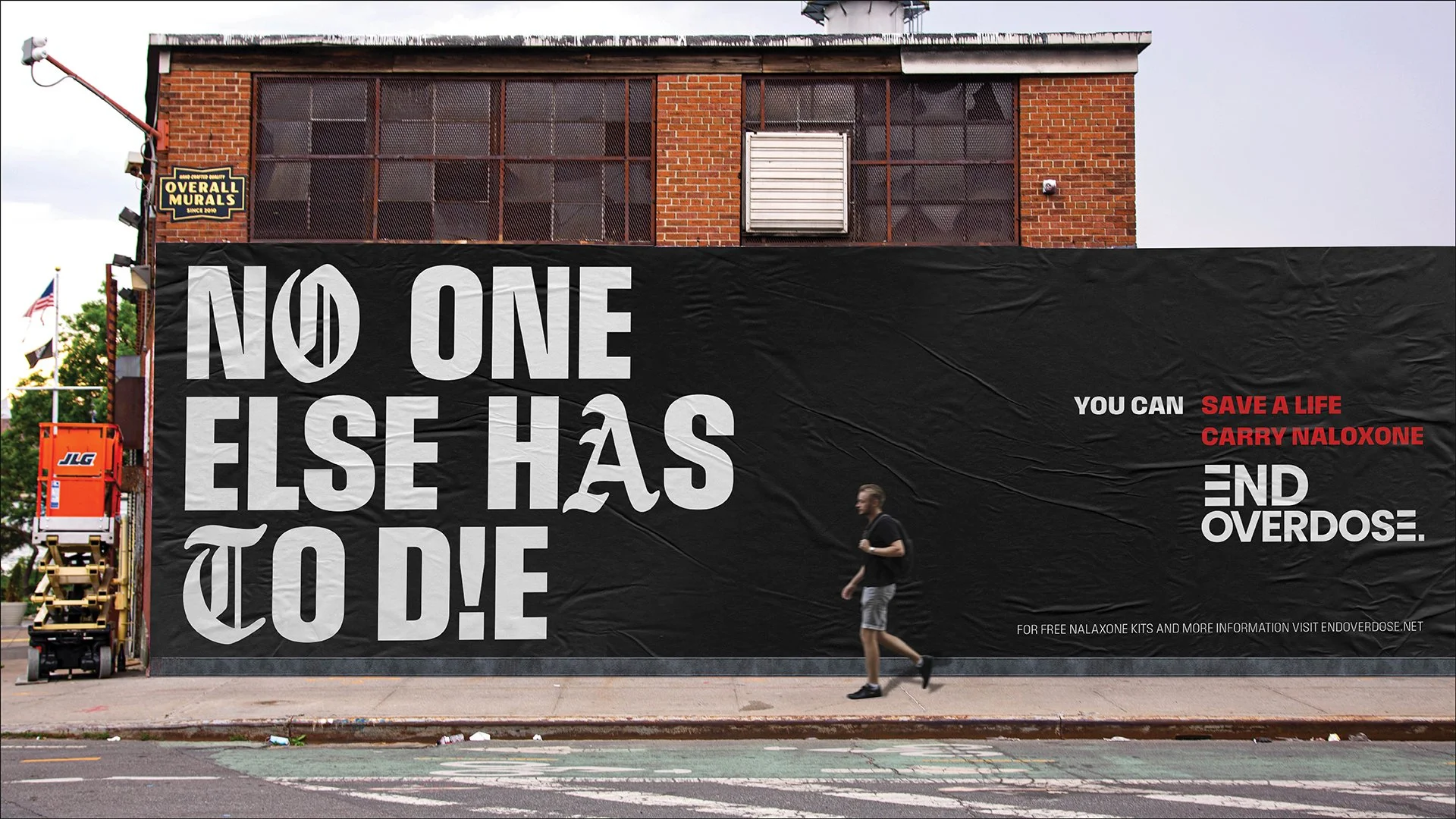 A large black banner on a building wall with white and red text providing an overdose awareness message about naloxone and overdose prevention. A person is walking past on the sidewalk.