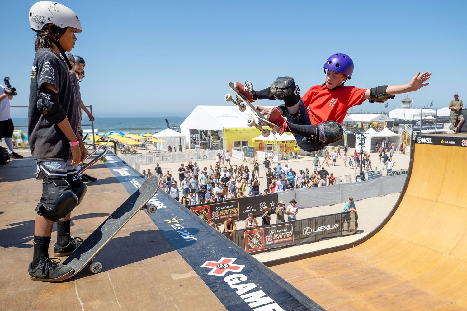 Skateboarder in red shirt and purple helmet launching off a ramp while another skateboarder watches at a skate park near the beach, with a crowd and tents in the background.