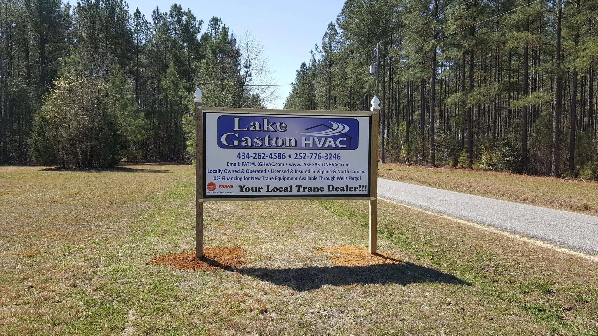 A large road side sign on 4x4 post outside the woods advertising Lake Gaston HVAC, with contact details and a logo, on a grassy area near a narrow road.