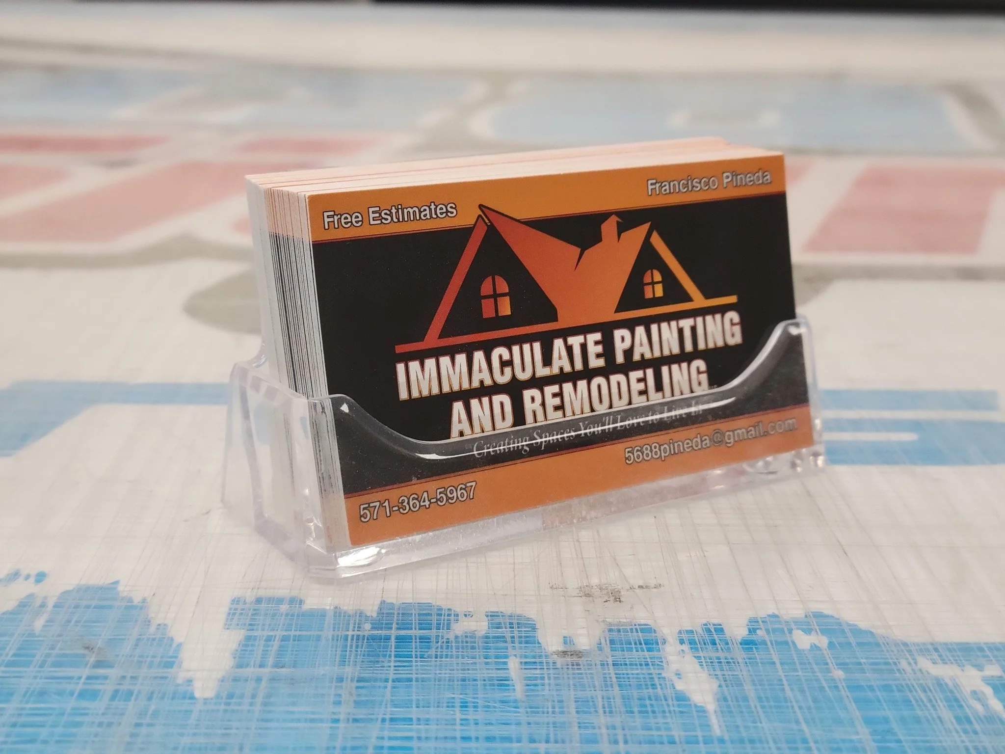 A business card holder with business cards promoting immaculate painting and remodeling services, including contact information and email address, on a table with a multicolored tablecloth.