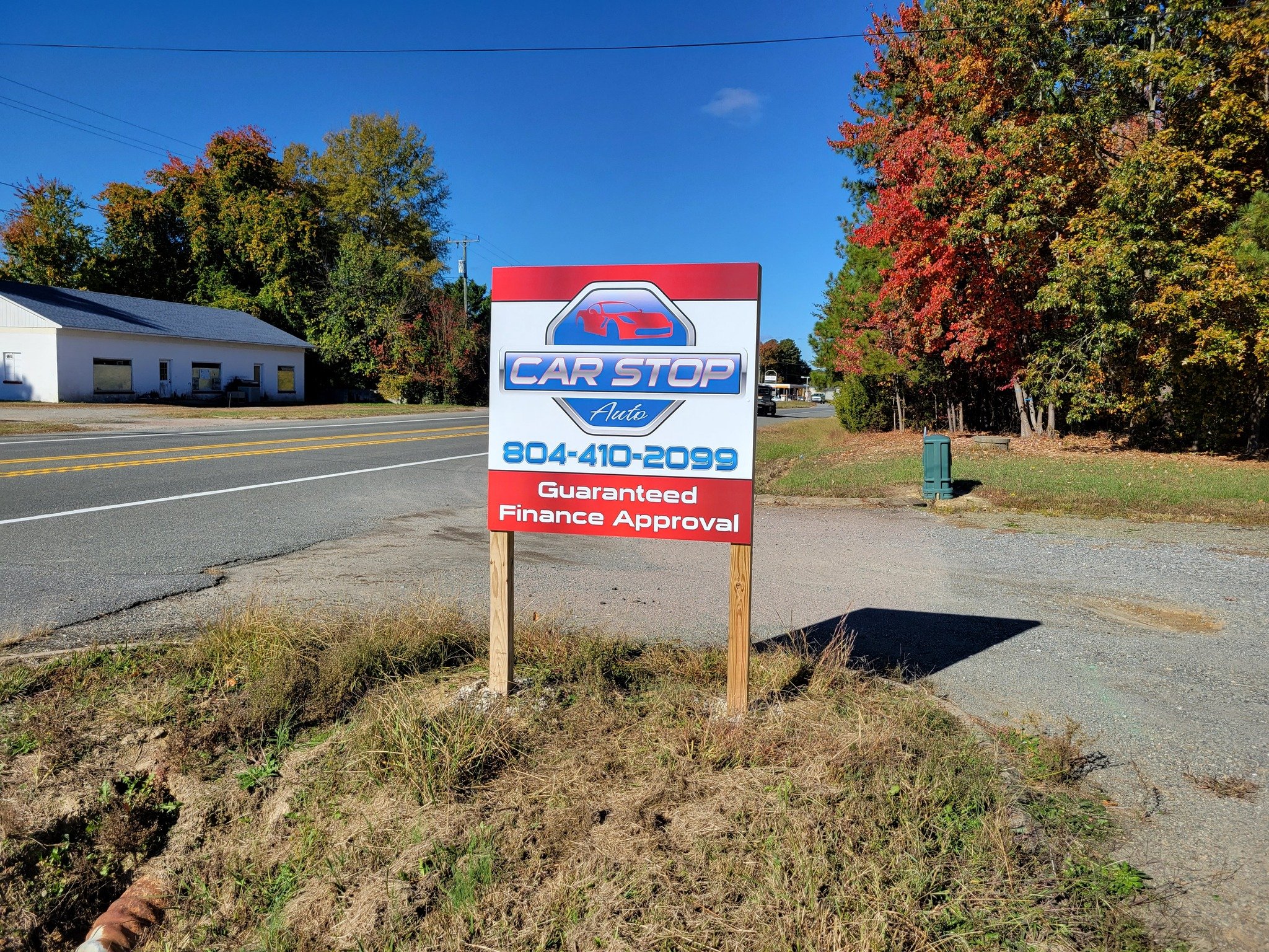 Custom designed signboard on roadside advertising Car Stop Auto, with phone number 804-410-2099 and guaranteed finance approval, situated in a rural area with trees and a building in the background under a clear blue sky.