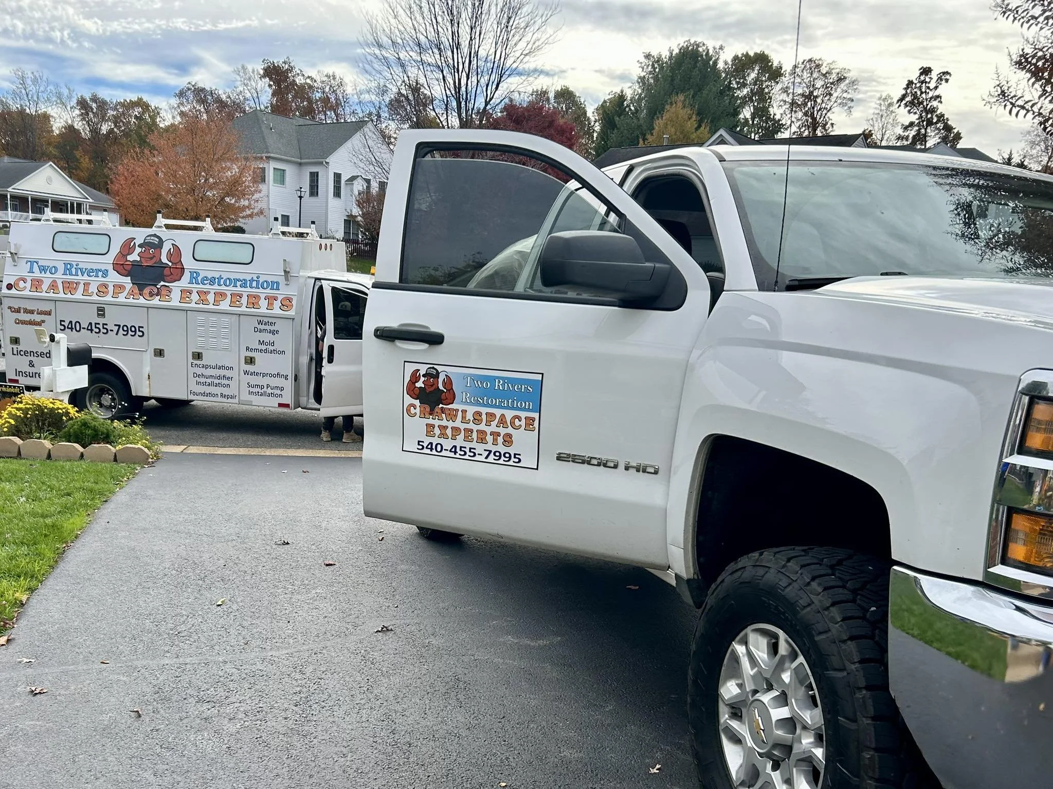 White pickup truck with vehicle magnets branded for Two Rivers Restoration parked on a driveway. Behind it, a van wrapped in vinyl graphics with similar branding is present. The background shows houses, trees with fall foliage, and a cloudy sky.