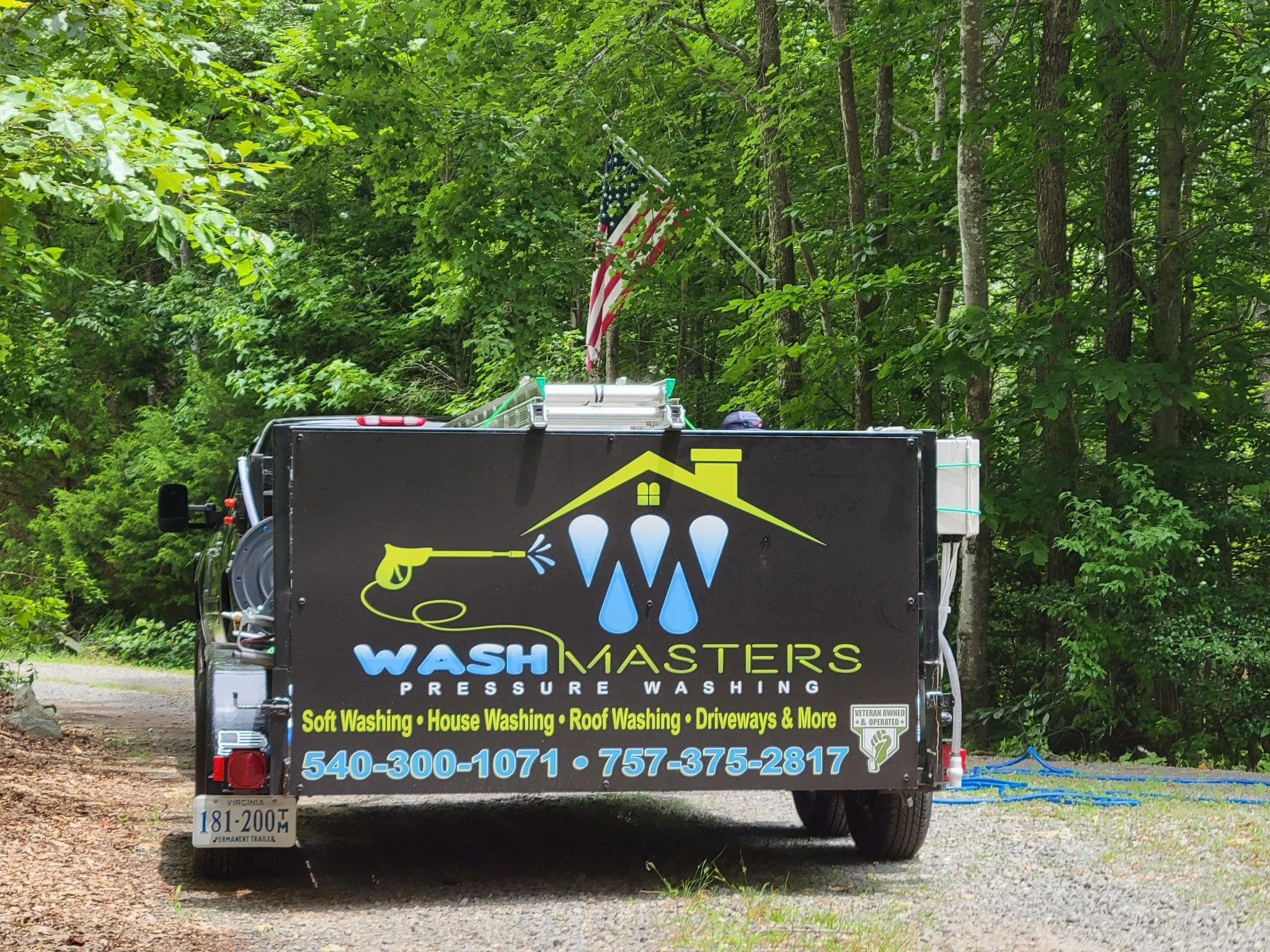 Aluminum sign mounted on a black utility trailer being pulled by a truck with the logo and contact information for Wash Masters, a pressure washing service, parked on a gravel area in a wooded area with green trees and a flag.
