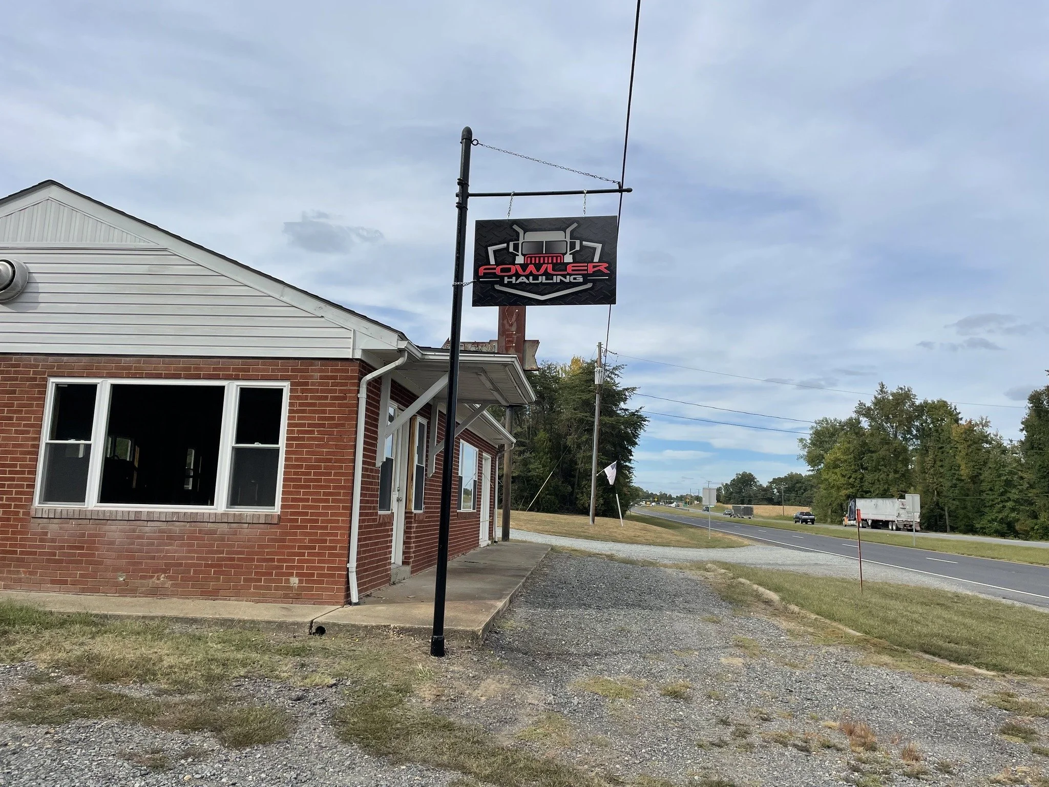 Hanging aluminum sign for Fowler Hauling next to a brick building on the side of a highway with trees in the background. The sign is branded and wrapped in vinyl graphics with the Fowler Hauling logo.