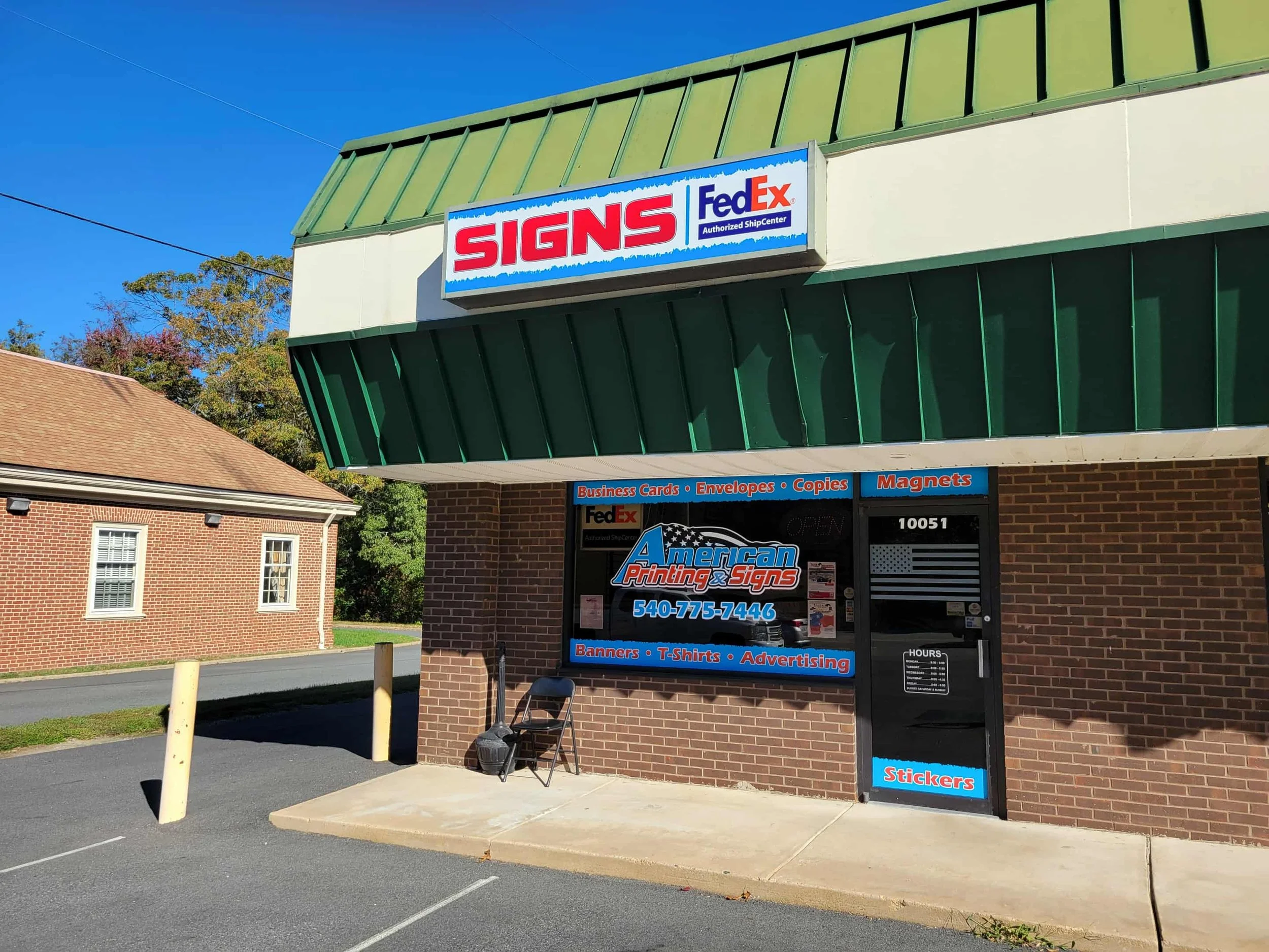 A storefront with a sign that reads 'SIGNS' and logos for FedEx, American Printing & Signs, with a list of services including business cards, envelopes, copies, magnets, banners, T-shirts, advertising, and stickers. The building has a brick exterior and a green metal awning, with a small sidewalk and parking lot in front.