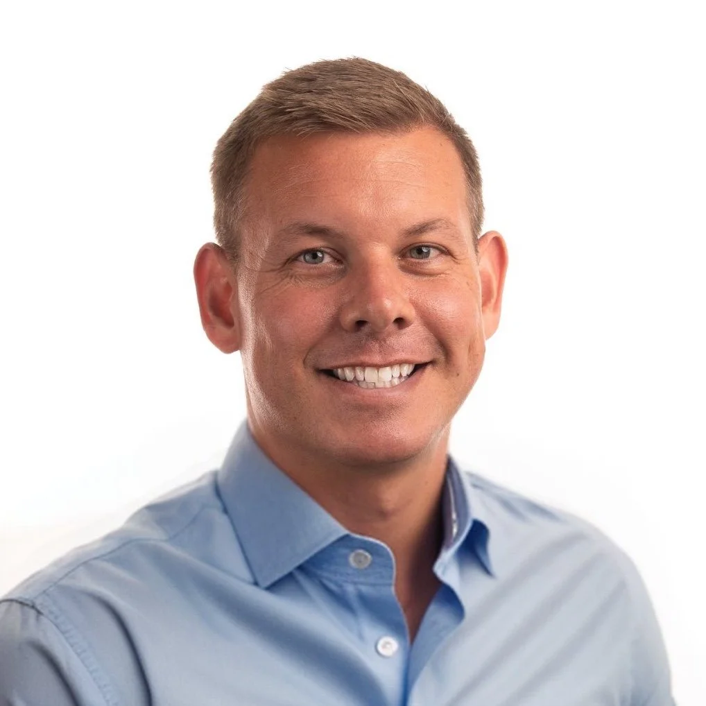 Portrait of a smiling man with short blonde hair wearing a light blue dress shirt against a white background.