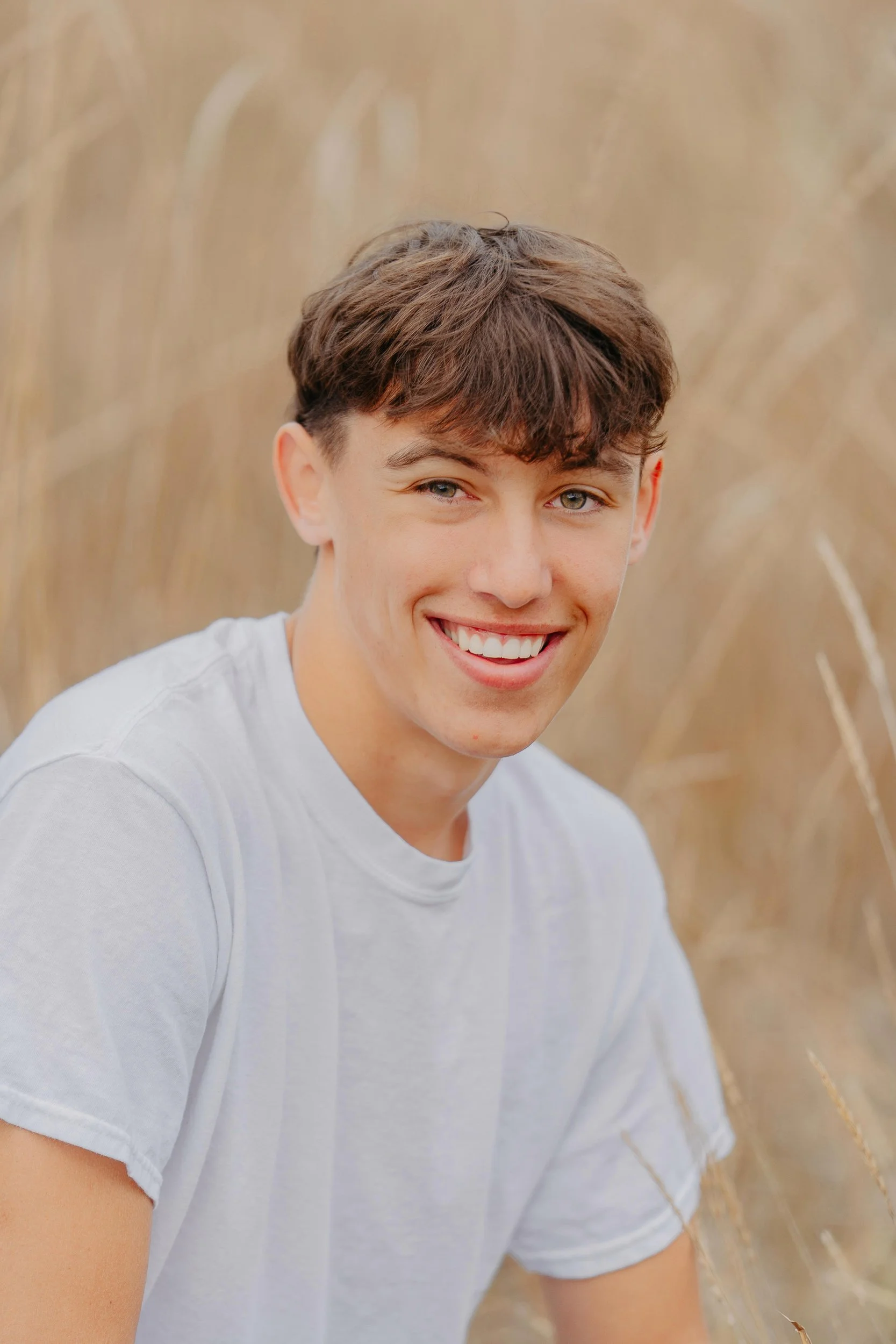 A young man with brown hair and blue eyes smiling while sitting outdoors in front of tall, dry grass.