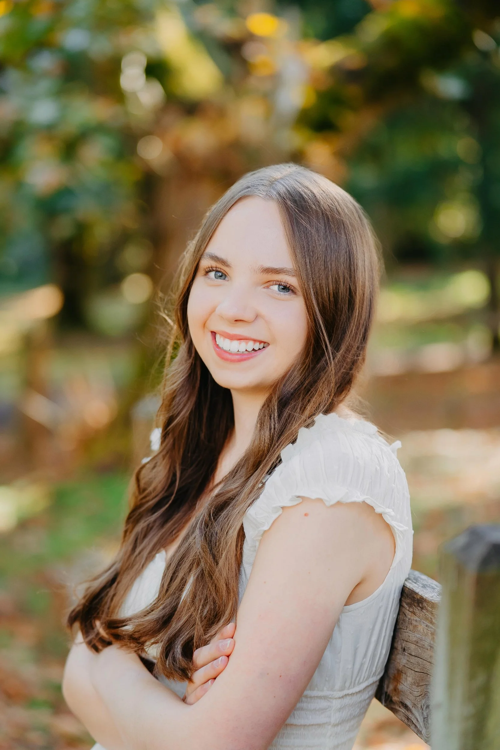 A young woman with long brown hair and blue eyes smiling outdoors in a park during autumn.