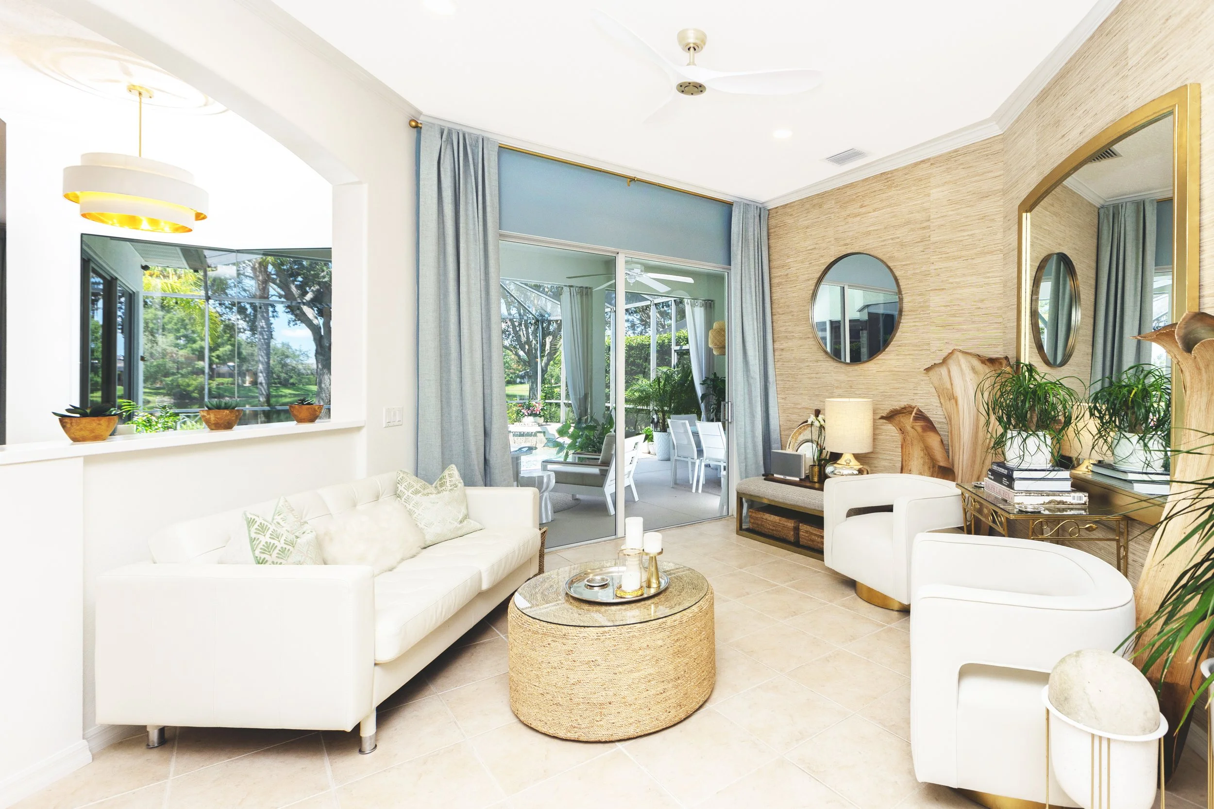 Living room with white sofas, a glass coffee table, decorative mirrors, potted plants, and a sliding glass door leading to an outdoor patio.