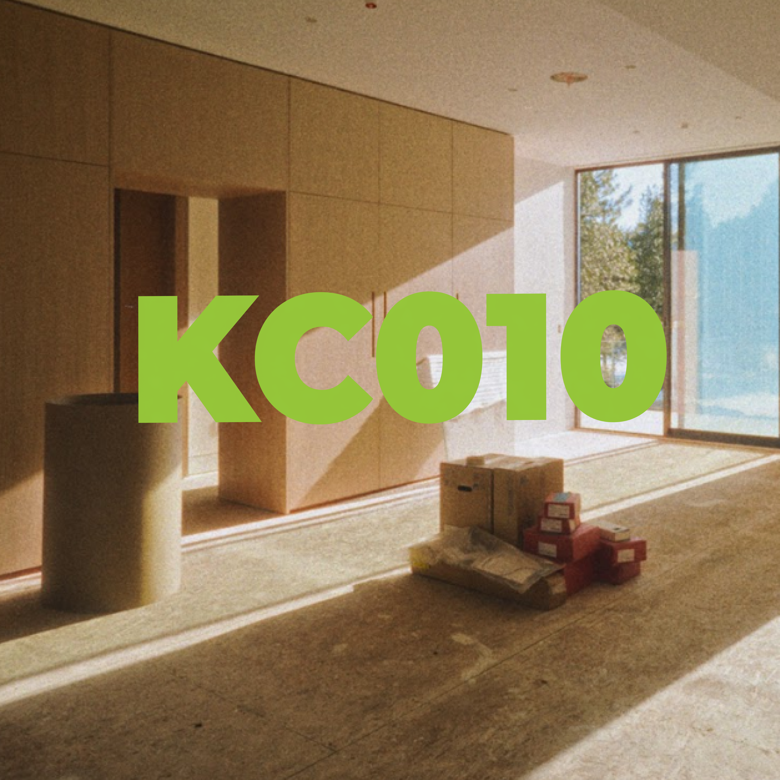 Interior view of an unfinished room with beige walls, large window with a view of trees, some boxes and construction materials on the floor, and the label 'KC010' in large green text.