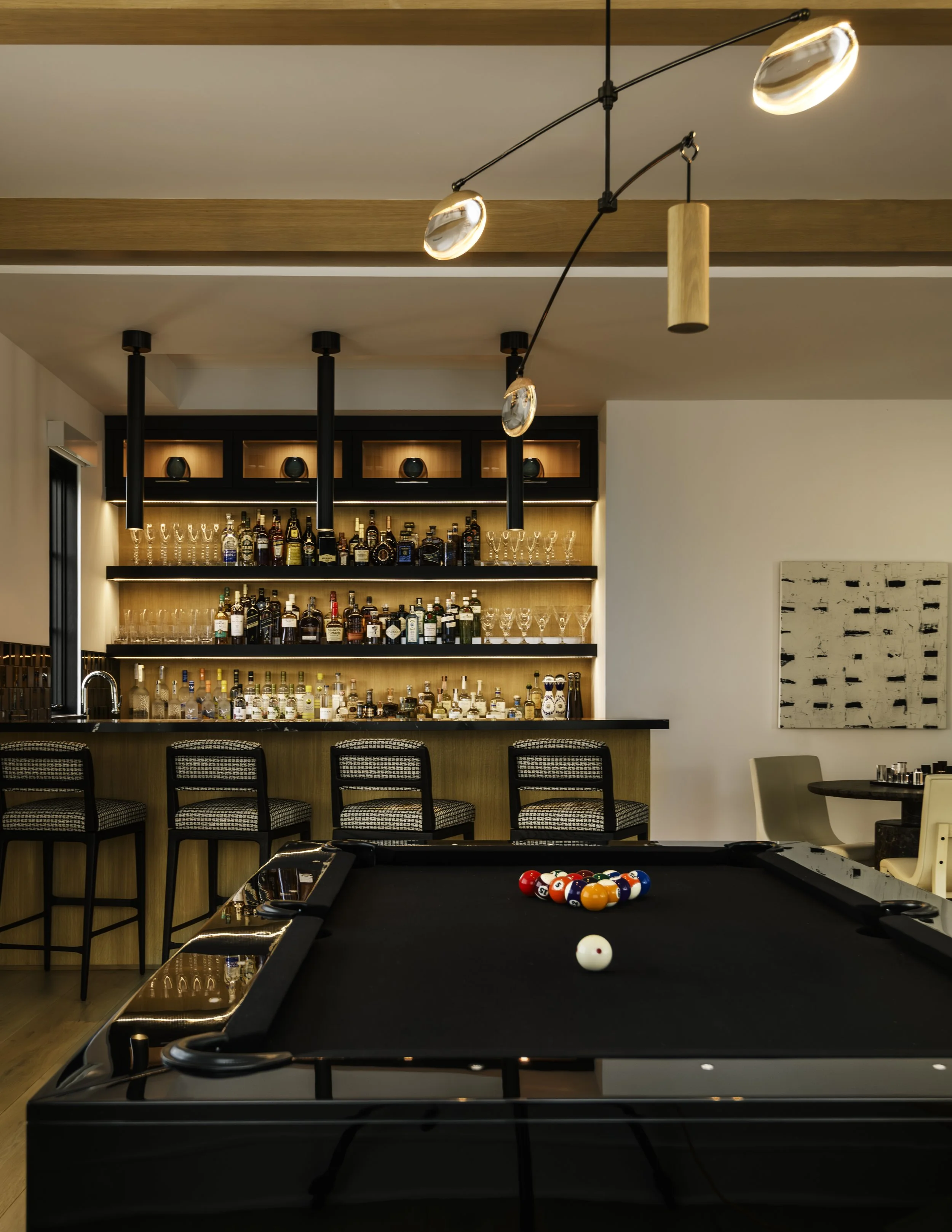 Indoor bar with a pool table in the foreground and a well-stocked bar behind it, with black bar stools and modern lighting fixtures.