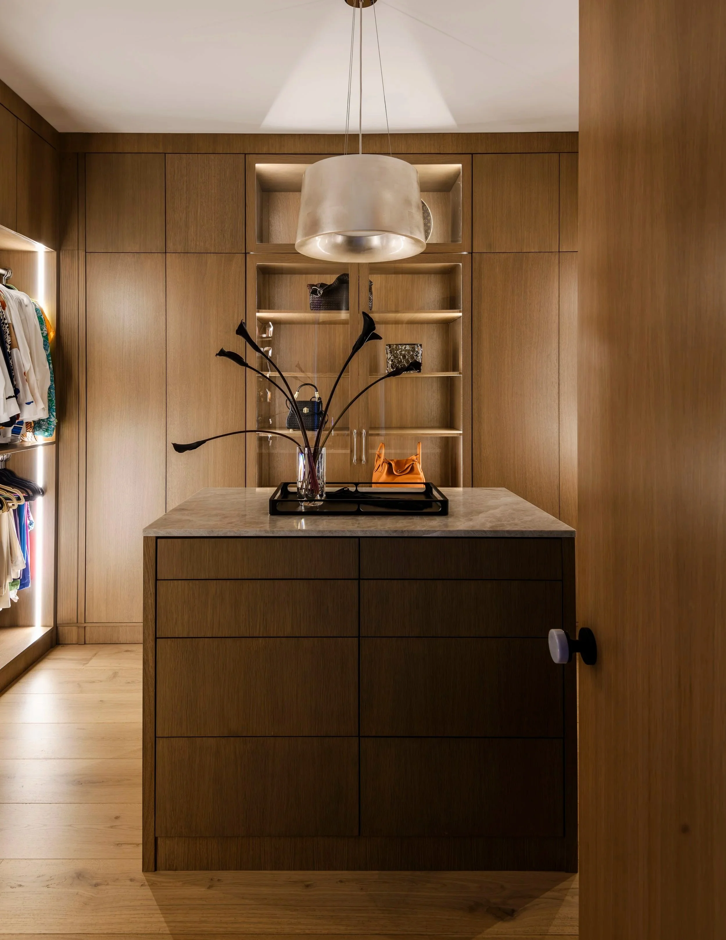 A modern walk-in closet with wooden cabinetry and a central island with a marble top. There is a decorative black vase with long-stemmed flowers on the island, along with a small orange handbag. Behind, open shelves displaying handbags and accessories. To the left, a wardrobe with clothes hanging is visible, and there's a large hanging light fixture above the island.