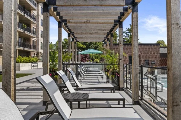 Outdoor patio with wooden pergola, loung chairs, sunshade umbrella, and surrounding greenery on a sunny day.