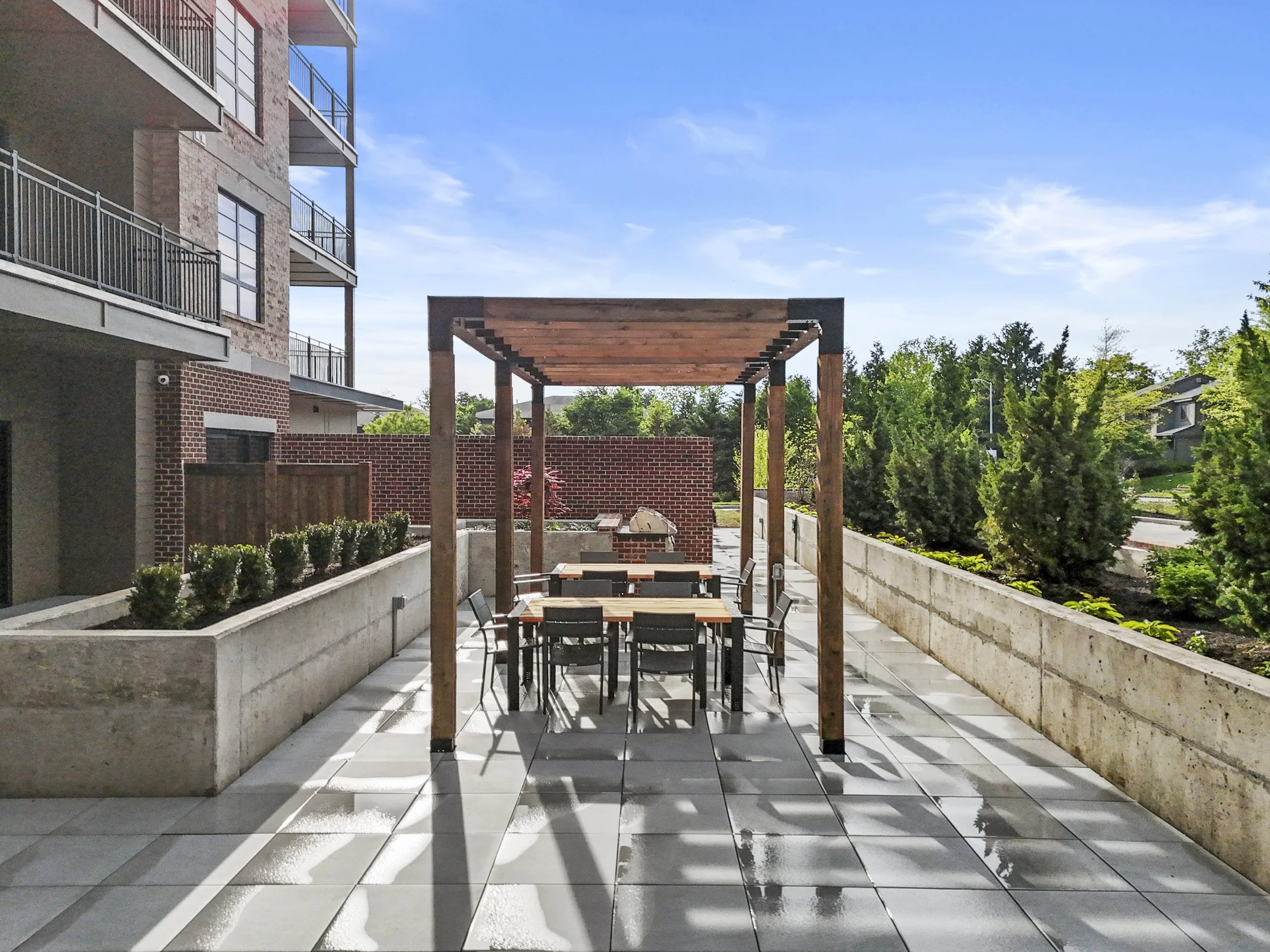 Outdoor patio with wooden pergola, dining table, black chairs, brick barbecue, and surrounding greenery on a sunny day.