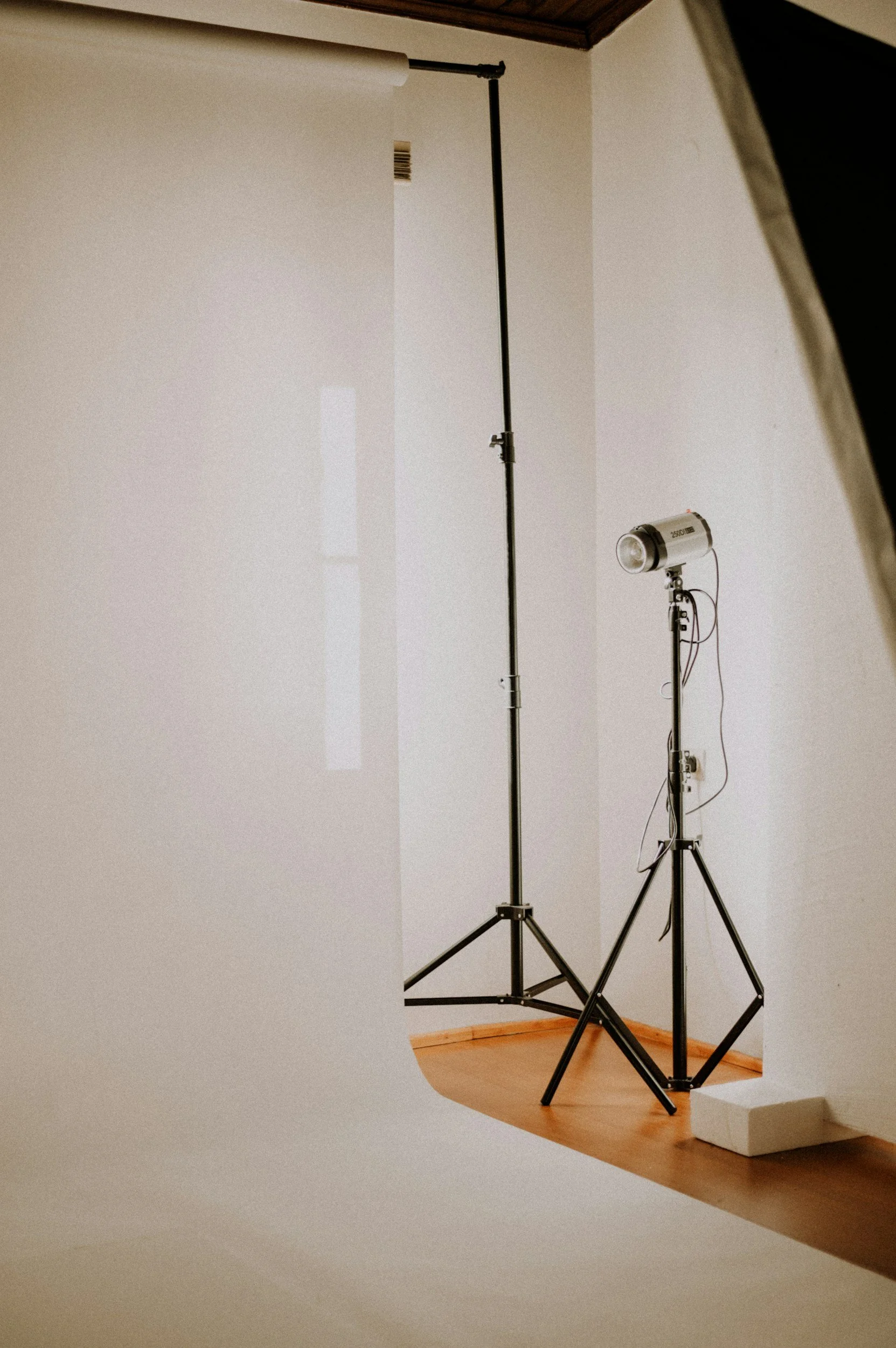 A photography set with white backdrop, two light stands, one with a spotlight, and a large roll of white paper unrolled on the floor. The setup is on a wooden floor in a room with a corner wall.
