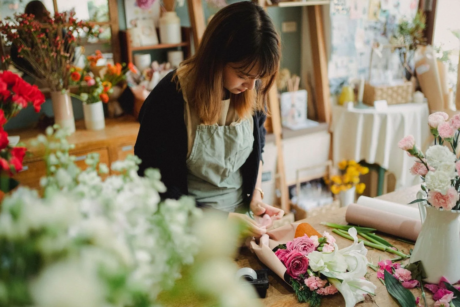 A woman working on floral arrangements in a flower shop. She is bending over a table with various flowers and wrapping floral foam.