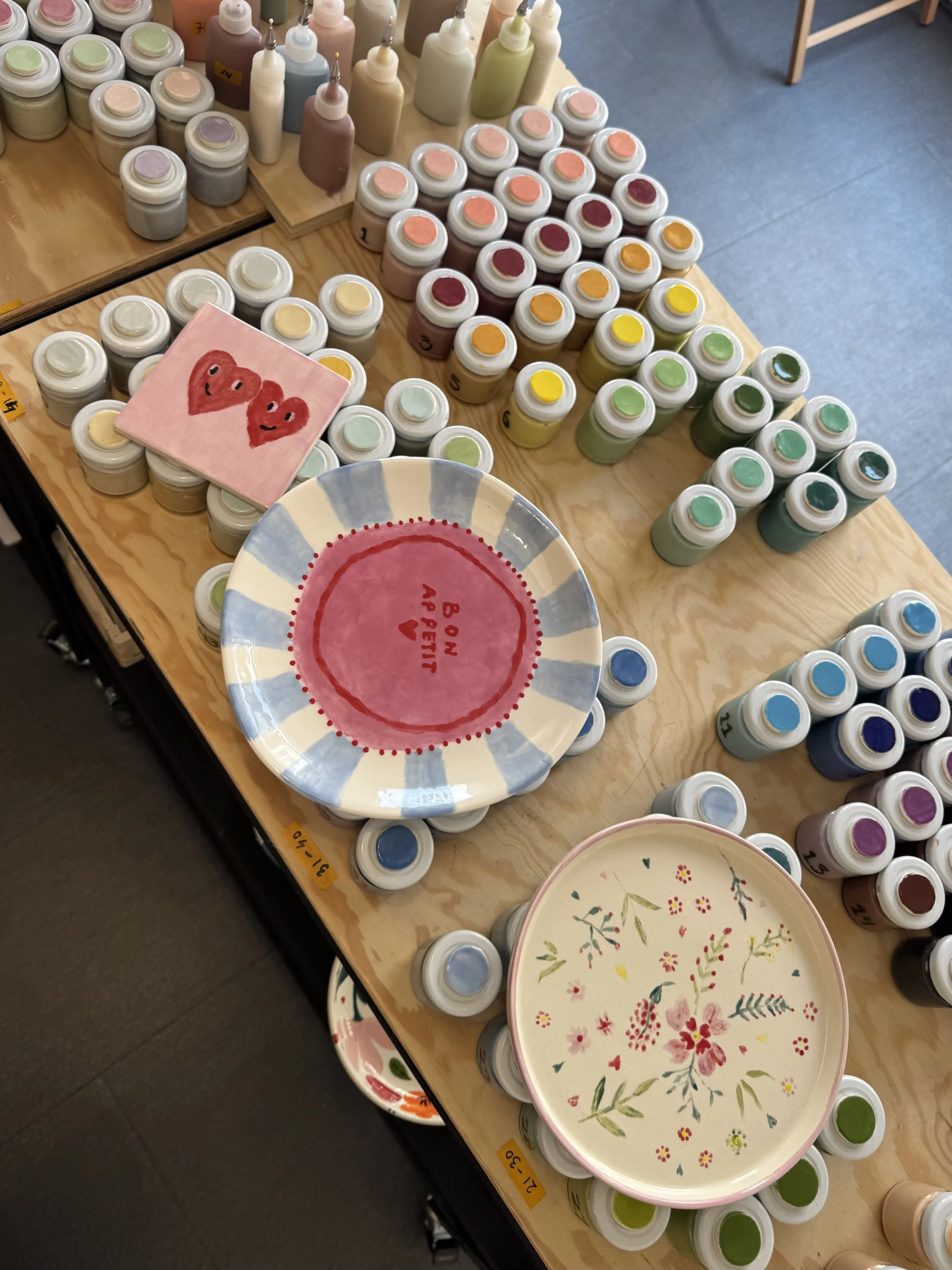 A display of small jars of pastel-colored paints, some bottles of liquid paint or dye, and decorative plates with floral patterns on a wooden table.