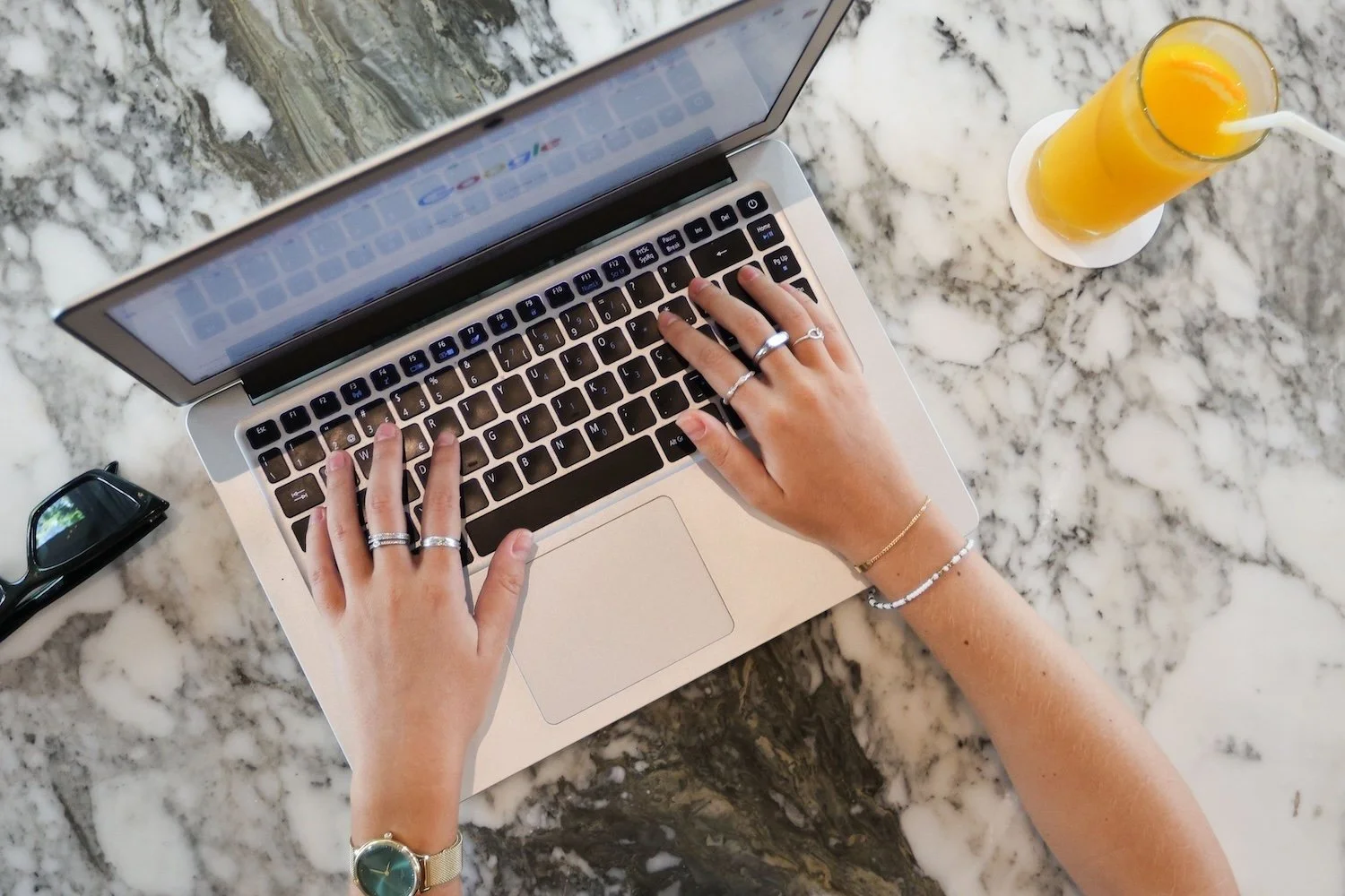 Hands typing on a laptop with a Google search open, next to a glass of orange juice on a marble table