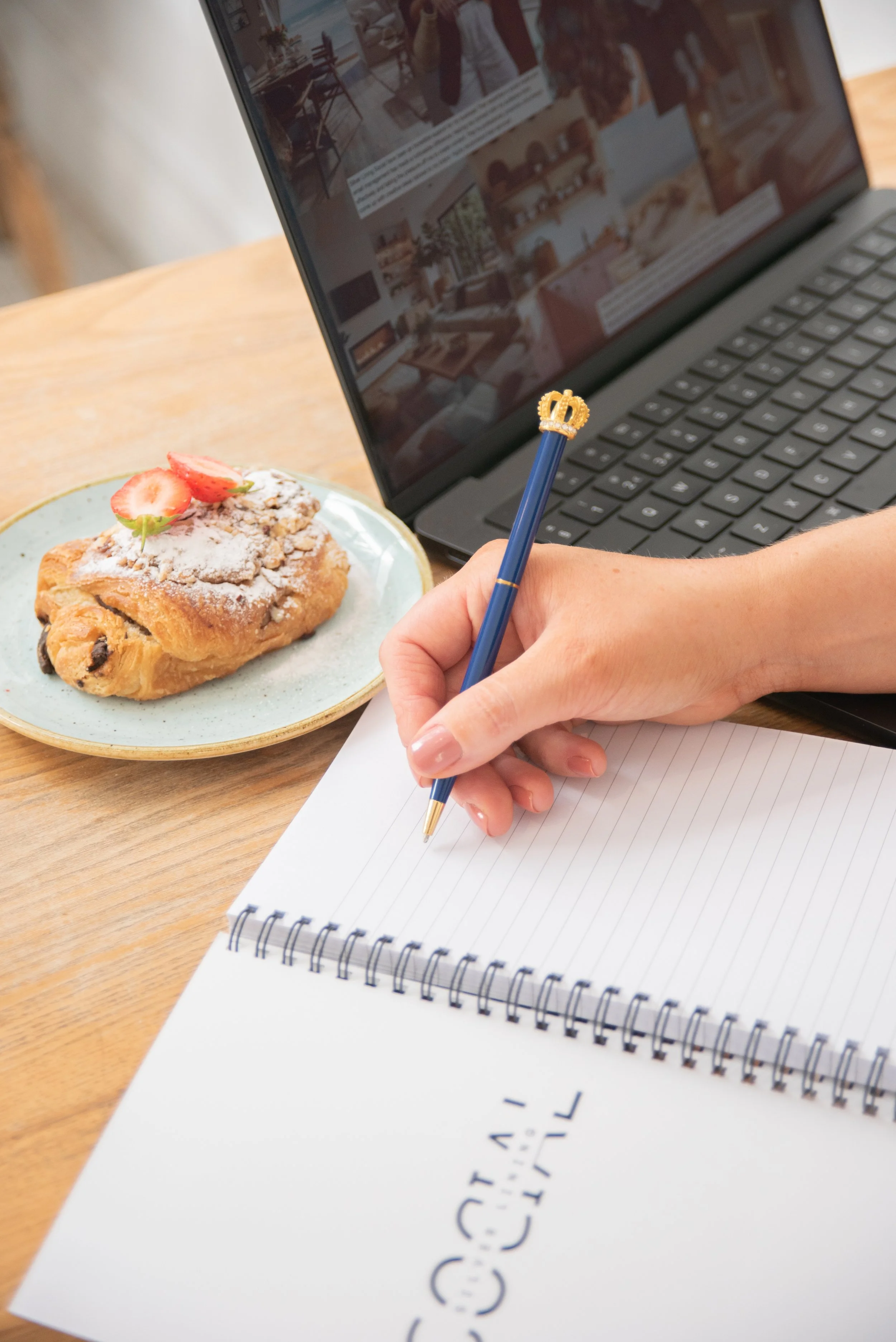 Content planning flatlay with a notebook, pen and pastry alongside a laptop displaying a website