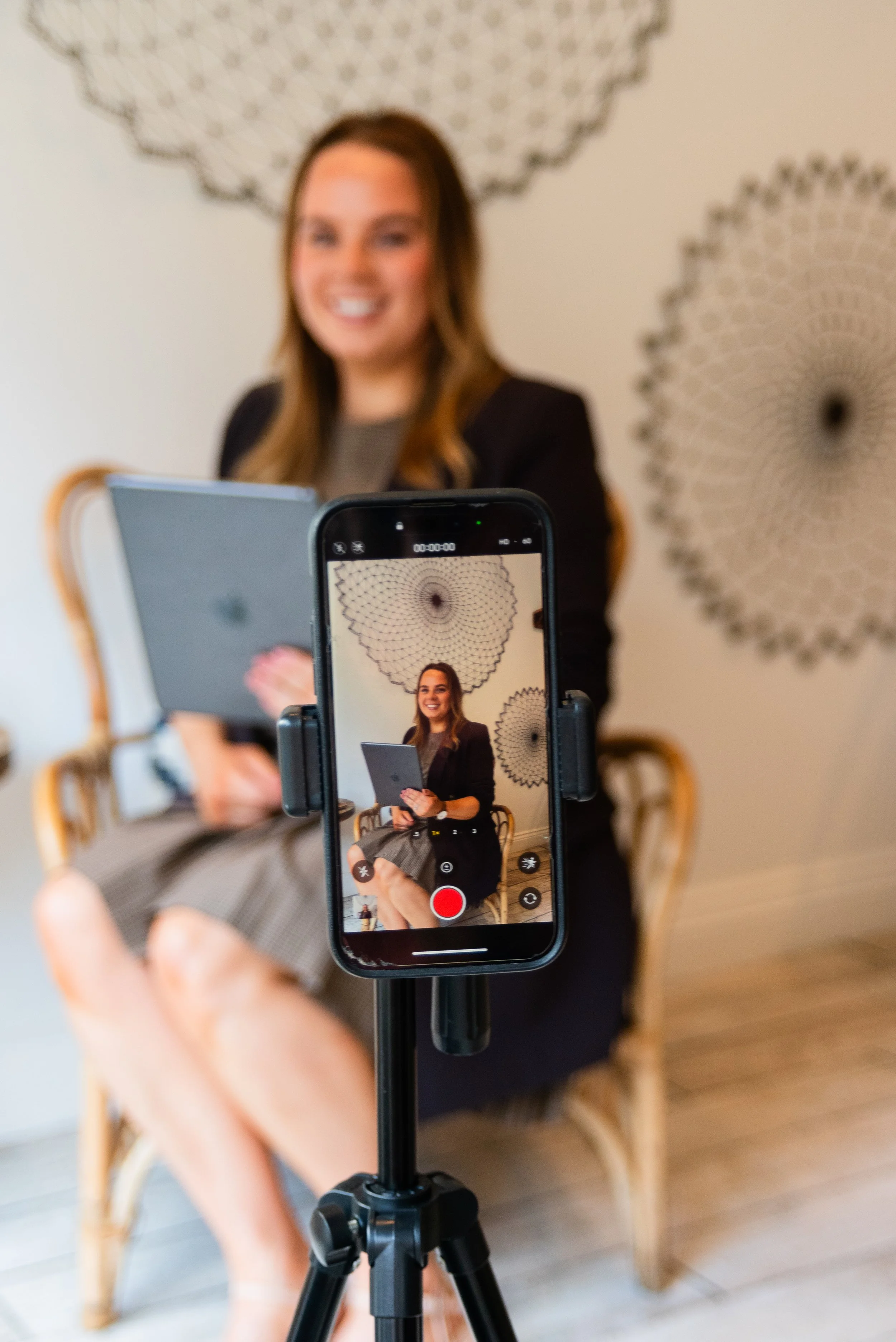 Grace Silver Lining Social sitting on a wooden chair, holding a tablet, is being recorded on a smartphone mounted on a tripod. The woman is smiling with a patterned wall in the background.