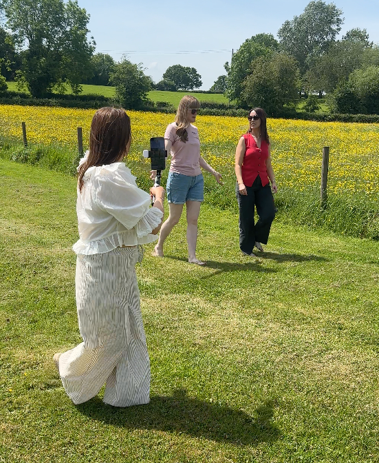 Content shoot in progress outdoors with a videographer filming two women walking and talking in a meadow
