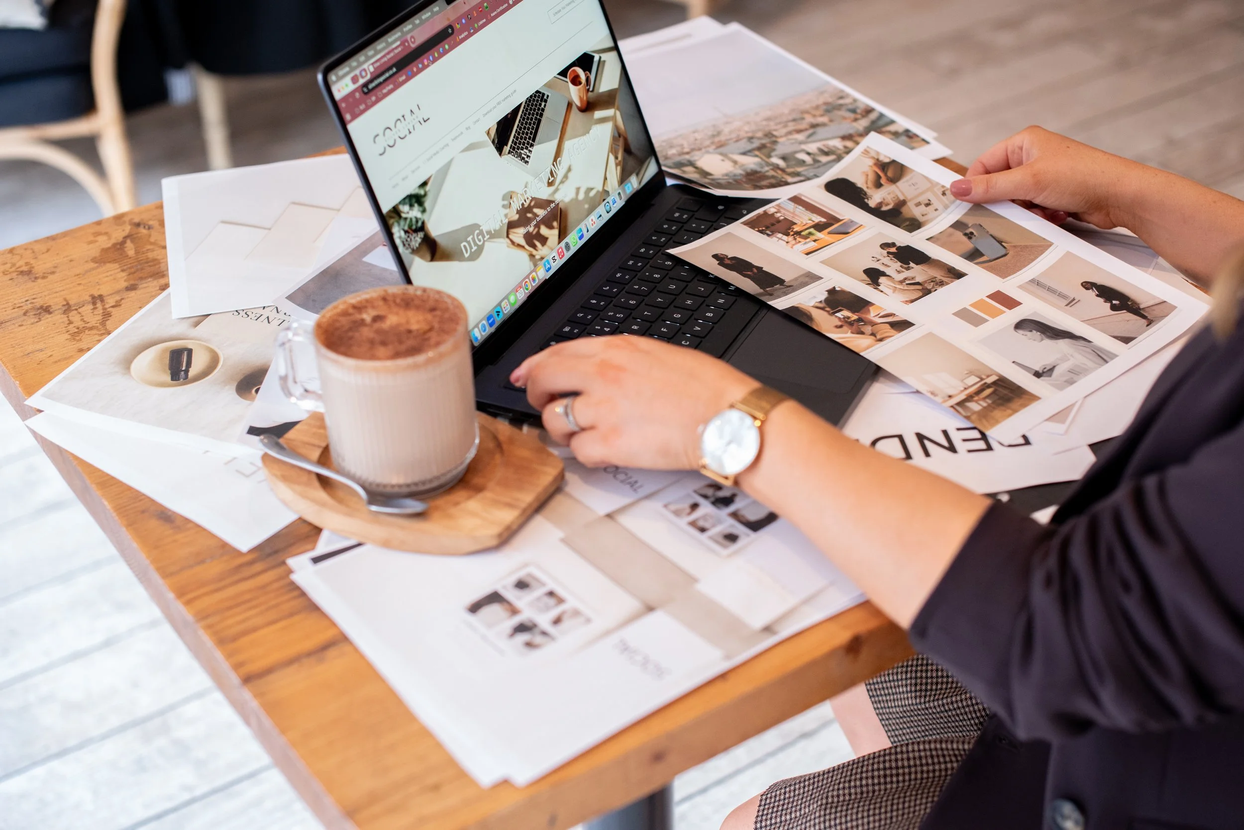 Grace Silver Lining Social working at a wooden table with a MacBook laptop, printed photographs, and a latte in a ceramic mug.