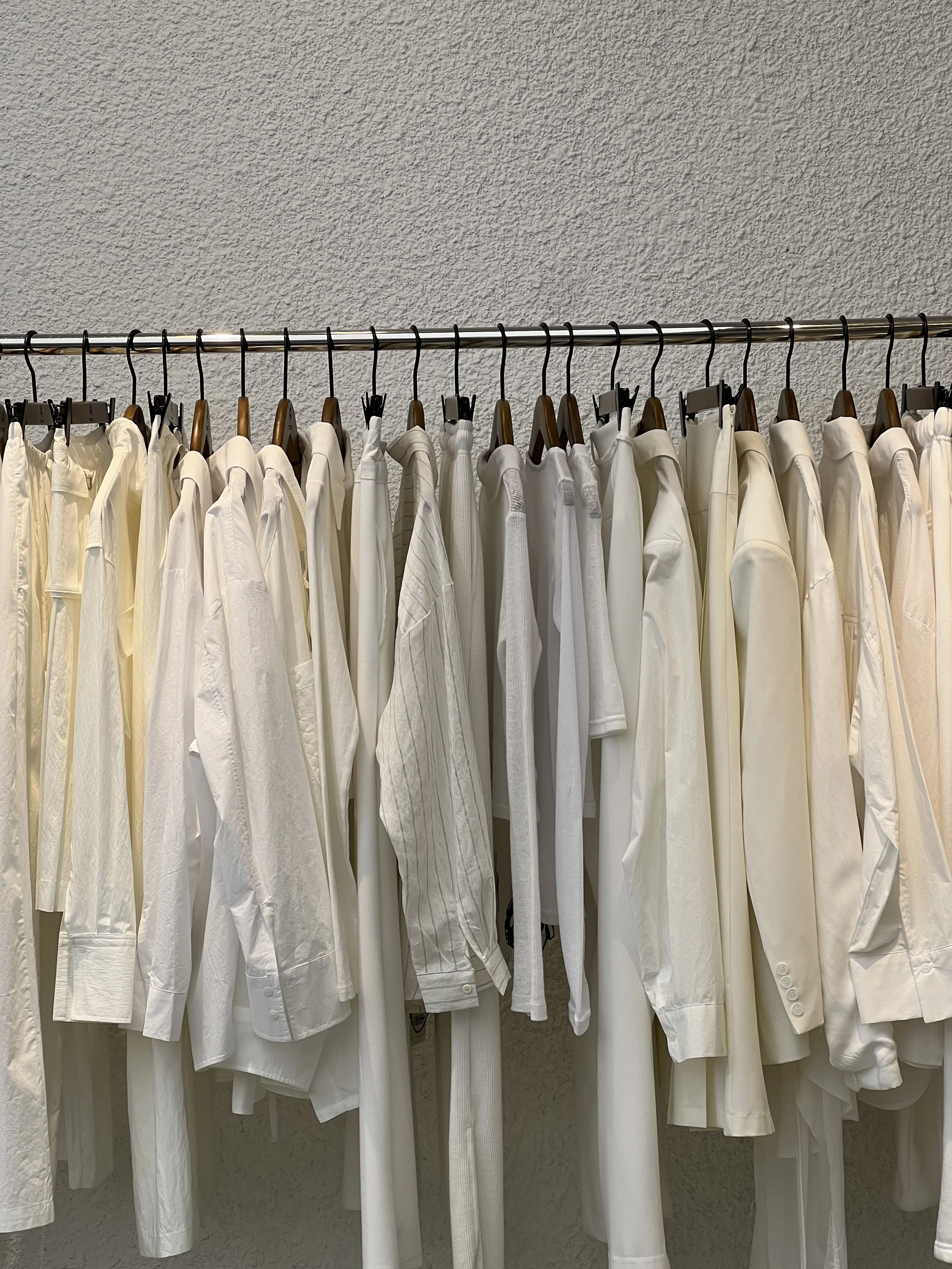 A clothing rack with white and cream-colored shirts and blouses hanging on wooden hangers against a textured beige wall.