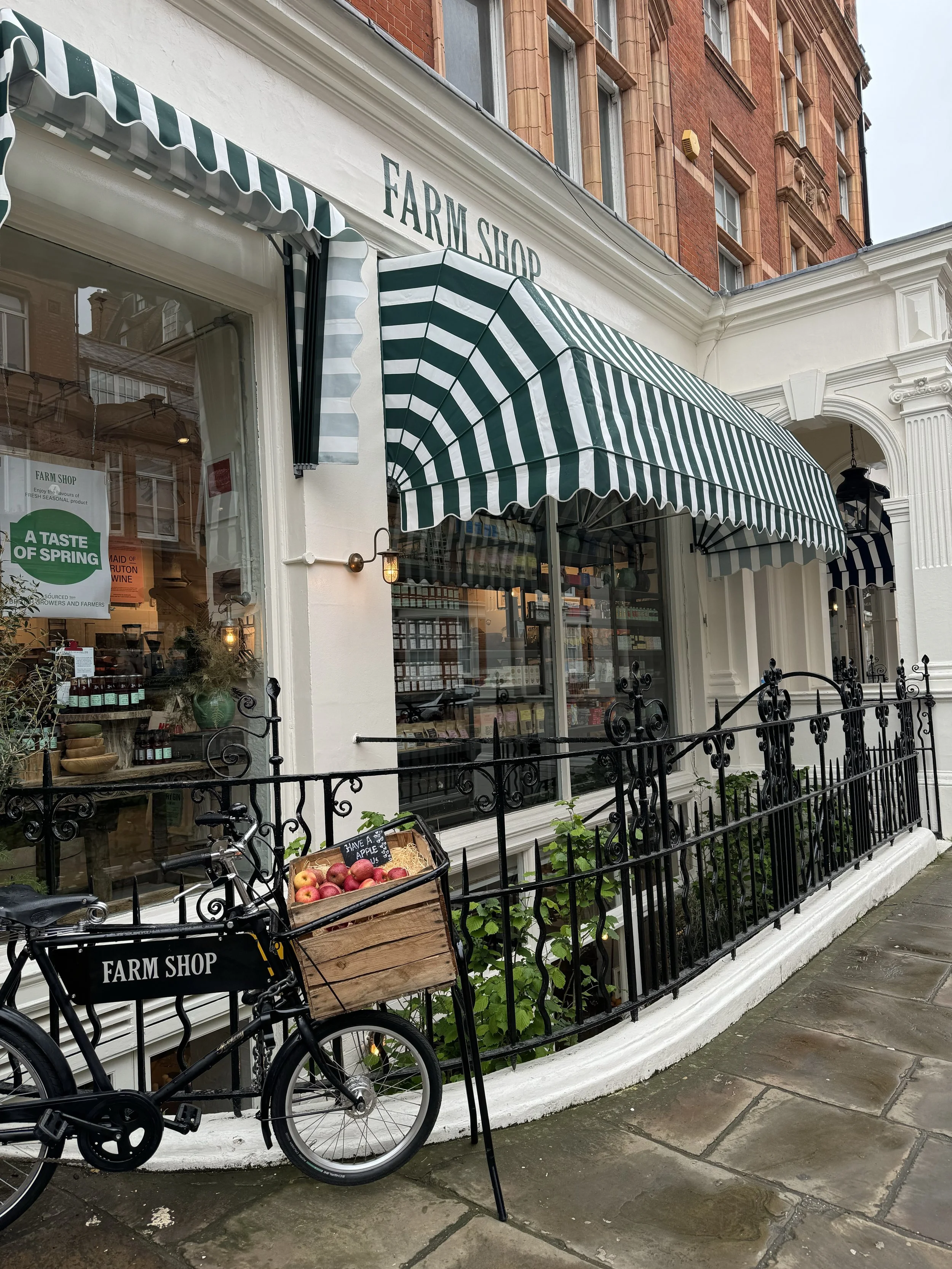 Exterior of a farm shop with green and white striped awnings, black decorative iron fence, and a bike with a basket of apples in front. The shop has large windows displaying products and signs, with a brick building above and a sidewalk in front.