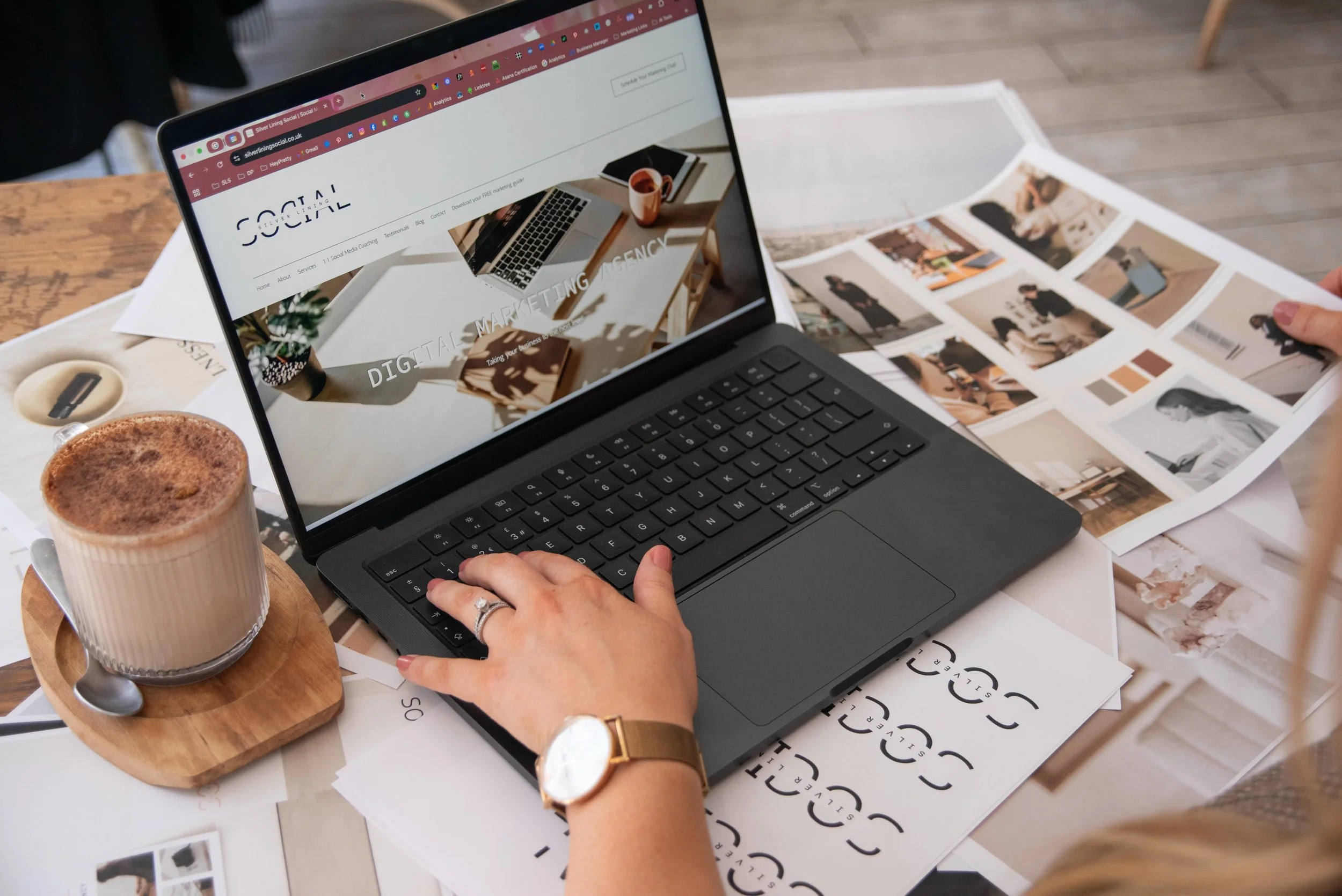 Hand working on a laptop displaying the Silver Lining Social website, surrounded by brand mood boards and a coffee on a wooden desk
