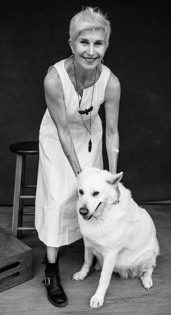 Bekah Vandenberg with short white hair smiling and wearing a white dress, standing next to a sitting white wolf, on a wooden floor with a dark background.