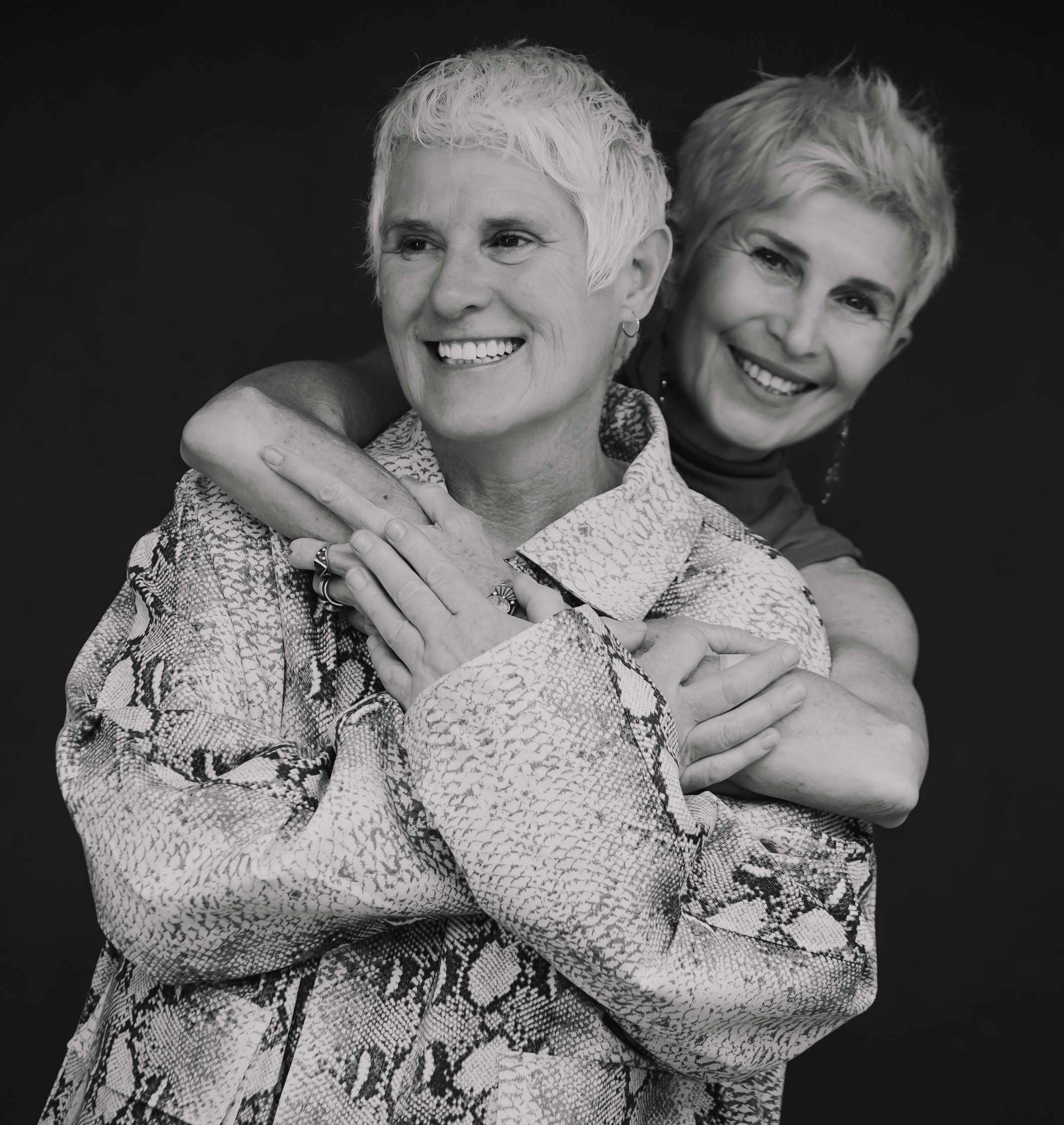 Black and white photo of Bekah Vandenberg and Cybele Rowe with short, light-colored hair, one embracing the other from behind against a dark background.