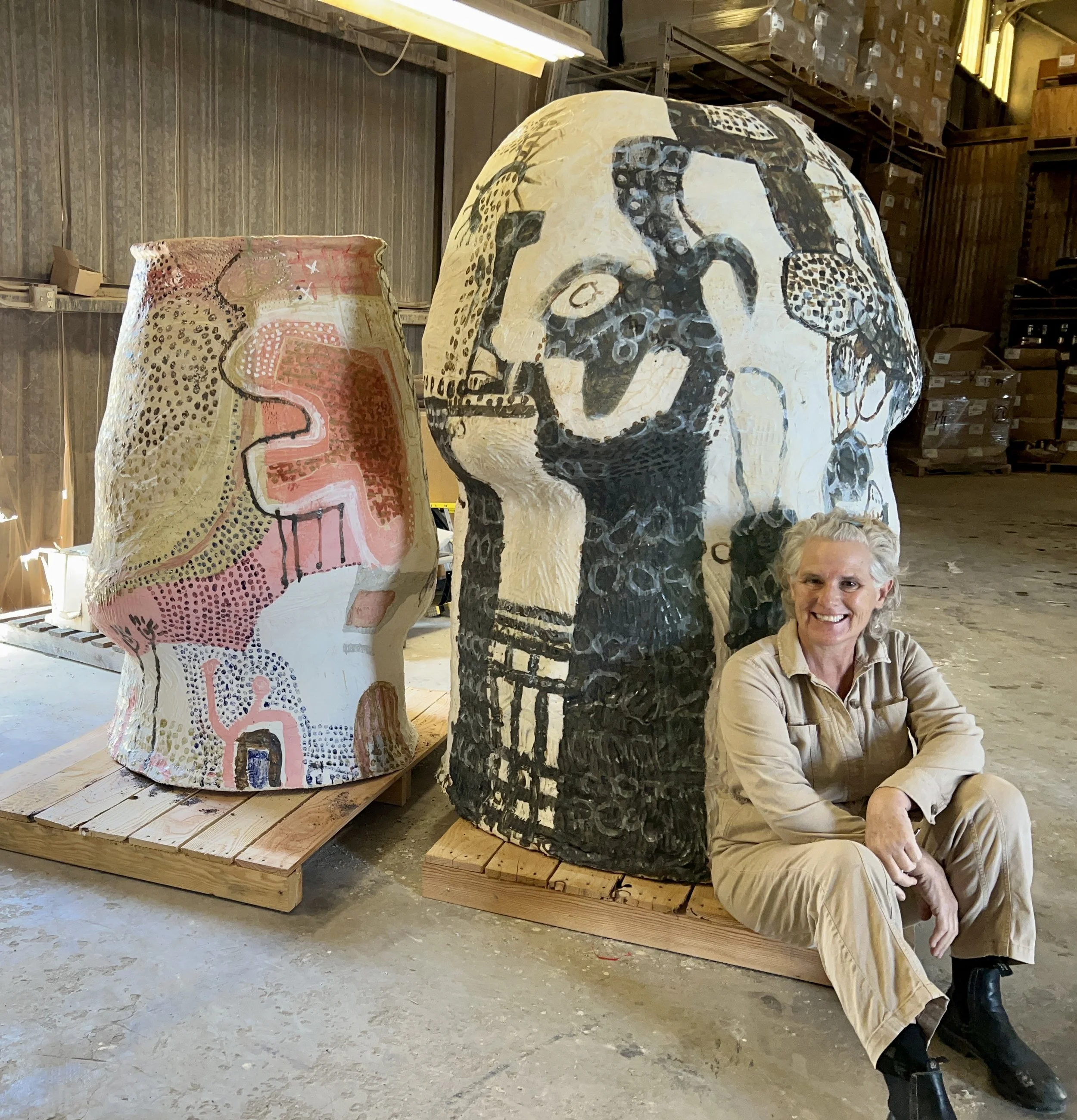 Cybele Rowe in beige clothing sits on the floor beside two large decorated sculptures in an industrial warehouse setting.