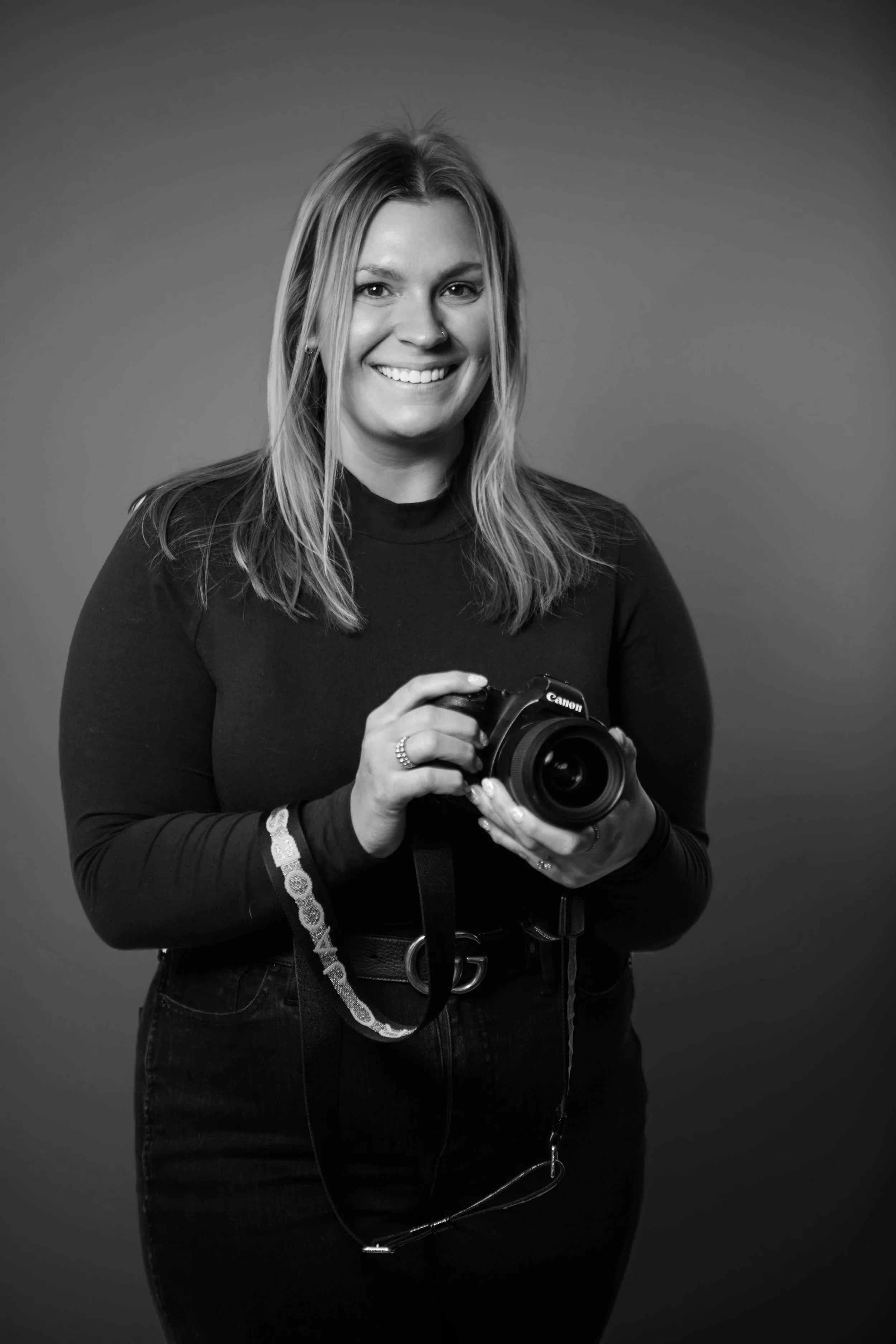 A woman with shoulder-length hair smiling and holding a professional camera in front of a plain background.