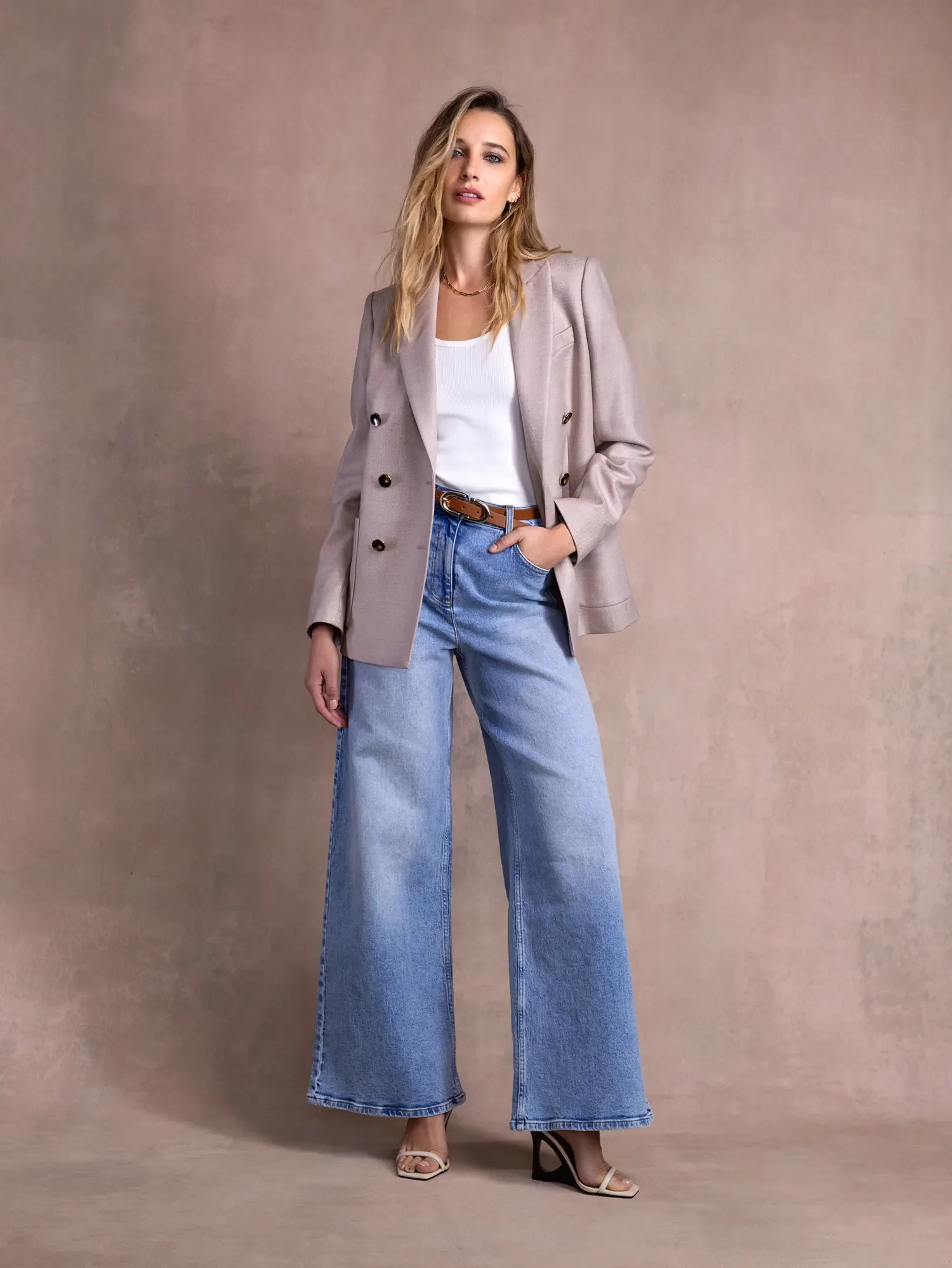 A woman with blonde hair wearing a beige blazer, white top, wide-leg blue jeans, and high heels standing against a beige background.