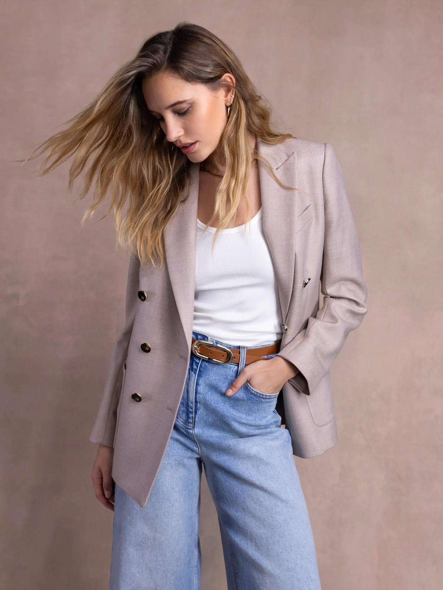 Woman with wavy hair wearing a beige blazer, white shirt, blue jeans, and a brown belt, standing against a plain background.