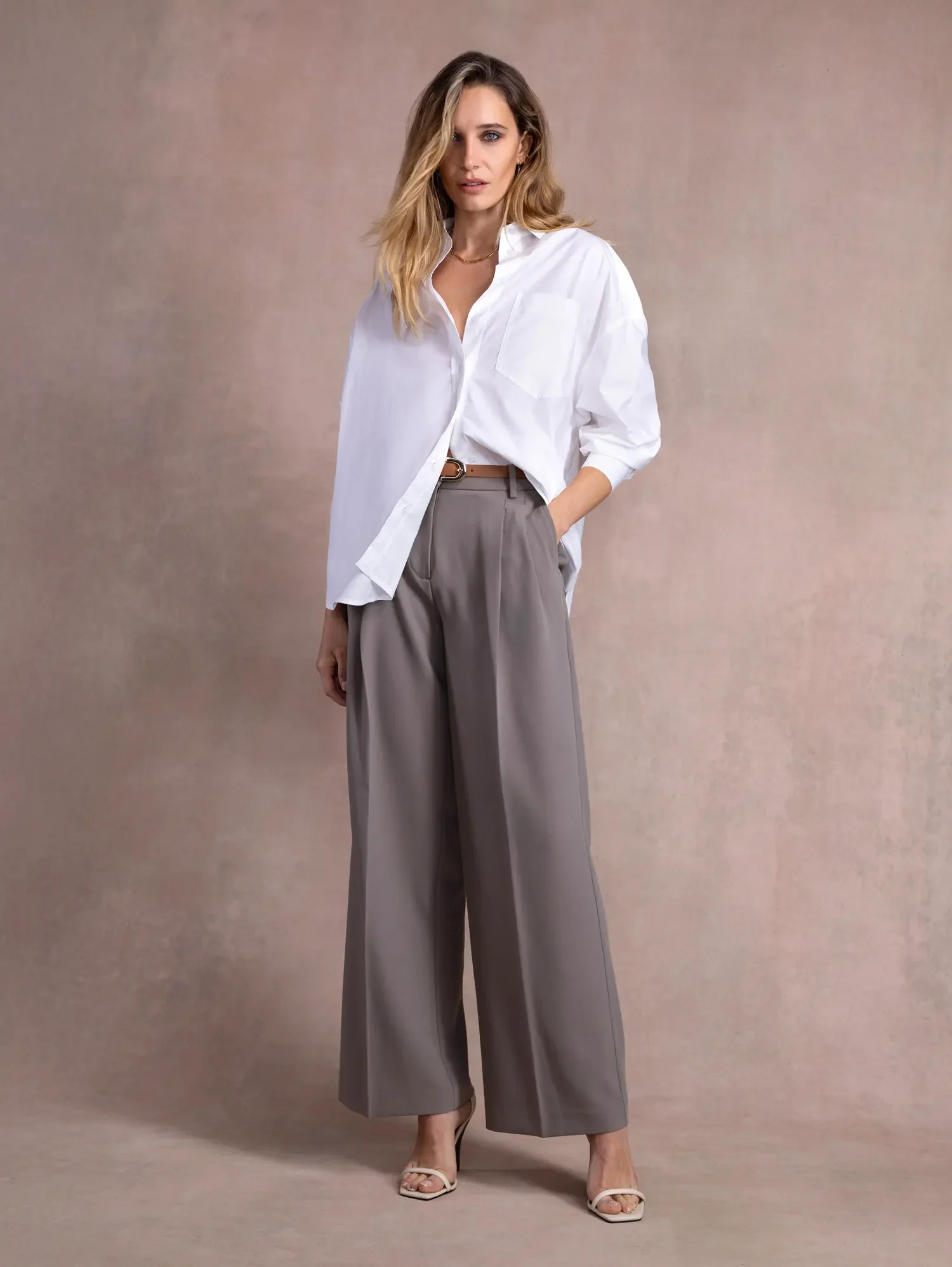 A woman with wavy blonde hair wearing a loose white button-up shirt, gray wide-leg pants, and white high-heeled sandals, standing against a neutral background.