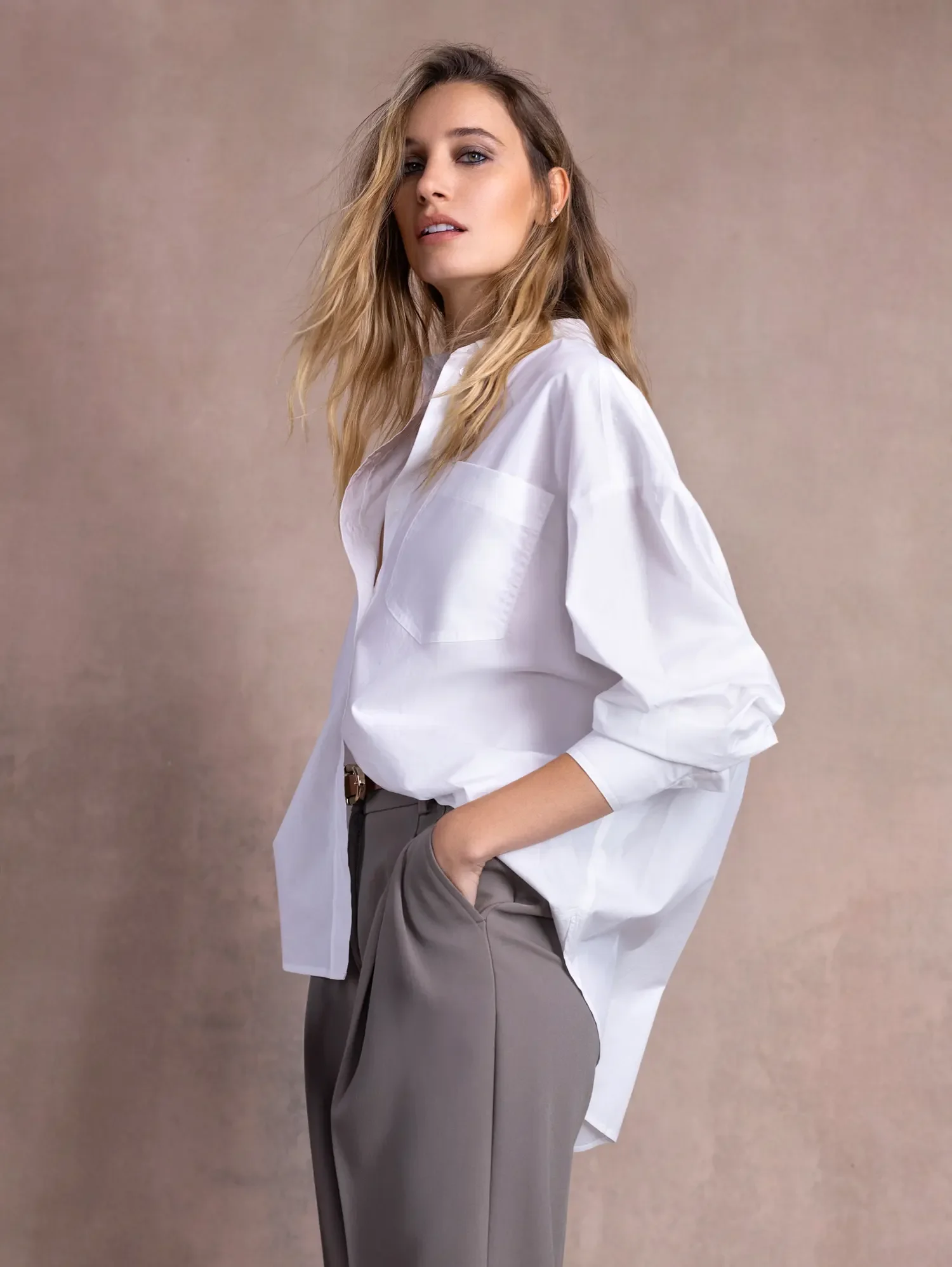 A woman with long wavy hair wearing a white button-up shirt and gray pants, standing against a neutral background, with one hand in her pocket looking at the camera.