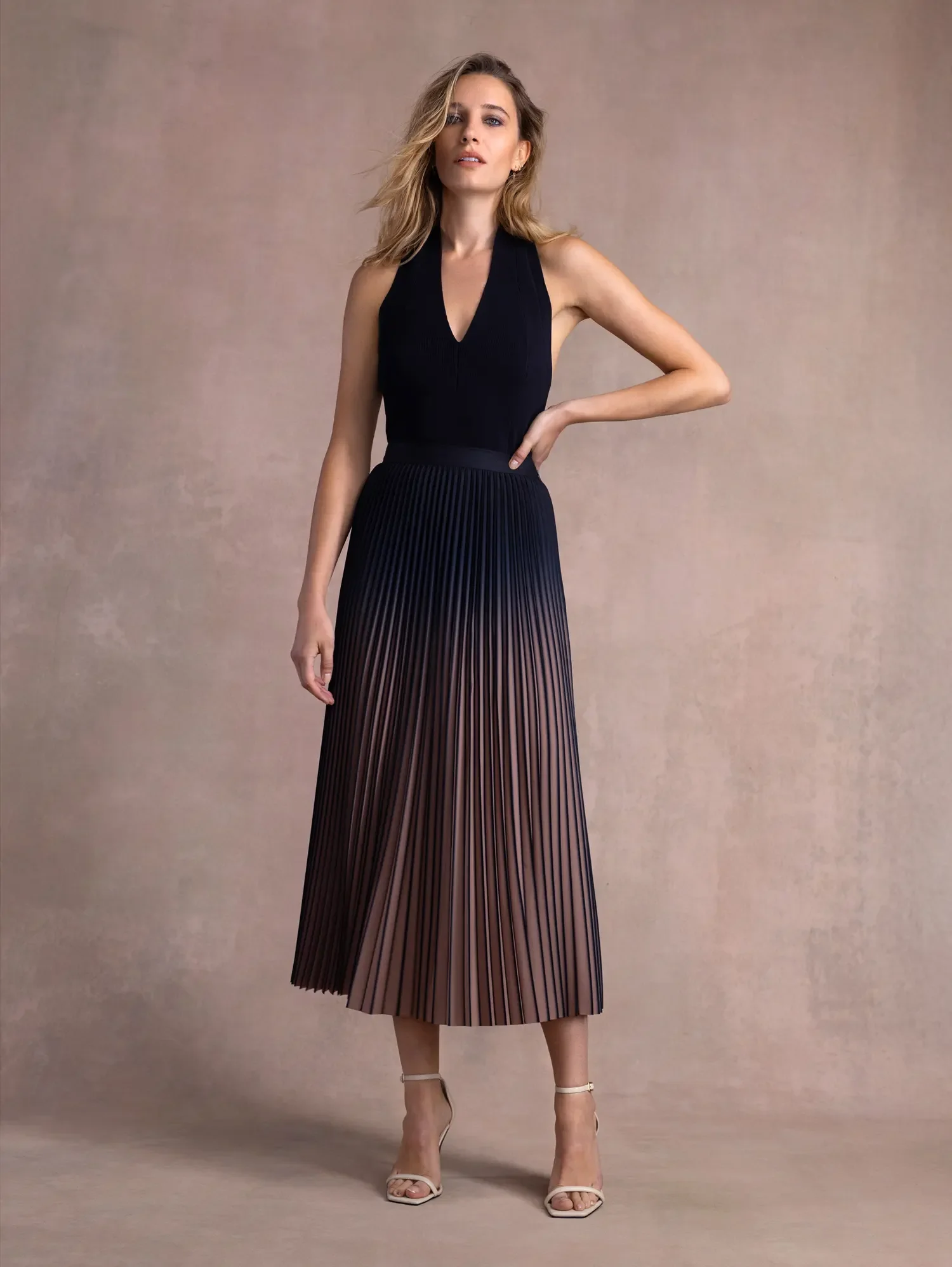 A woman in a black sleeveless V-neck top and a pleated, multicolored midi skirt stands against a plain beige background. She has blonde, wavy hair and wears beige high-heeled sandals.