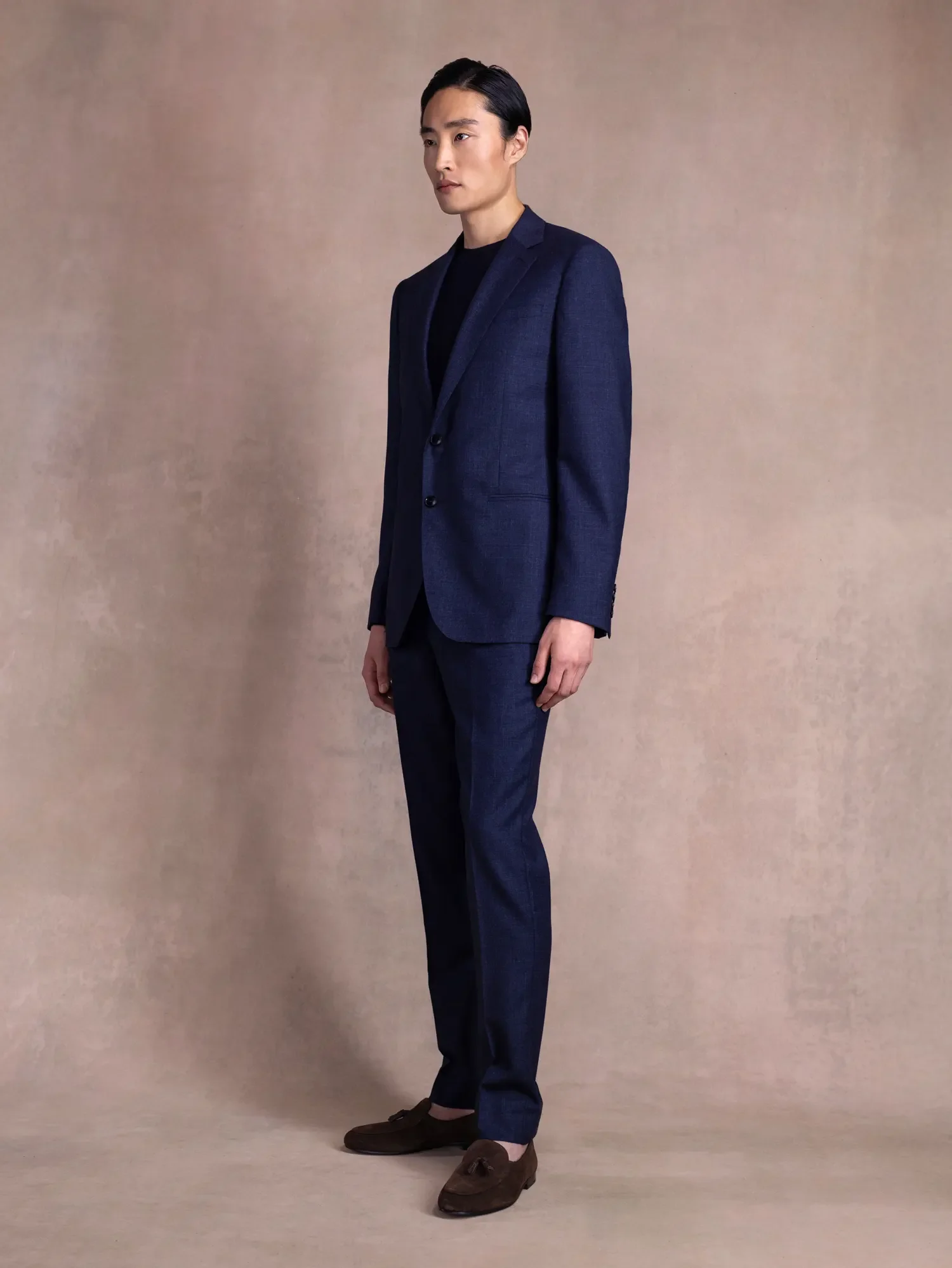 A man in a navy blue suit and black shirt standing against a plain beige background.