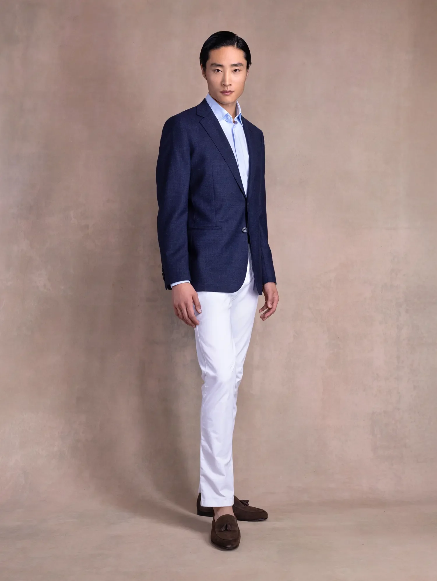 A man wearing a navy blue blazer, light blue shirt, white pants, and brown shoes standing against a beige background.
