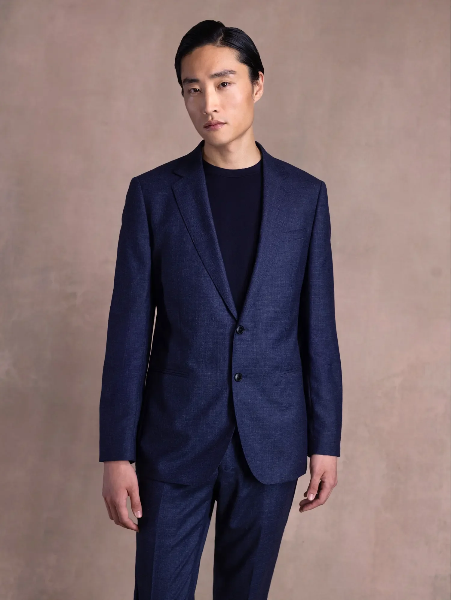 A person wearing a dark blue suit, black shirt, and standing against a plain beige background.