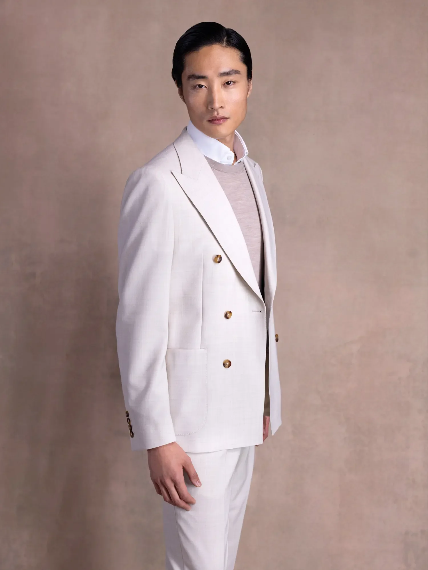 A man with black hair in a white suit, layered over a beige sweater and white button-up shirt, standing against a beige background.